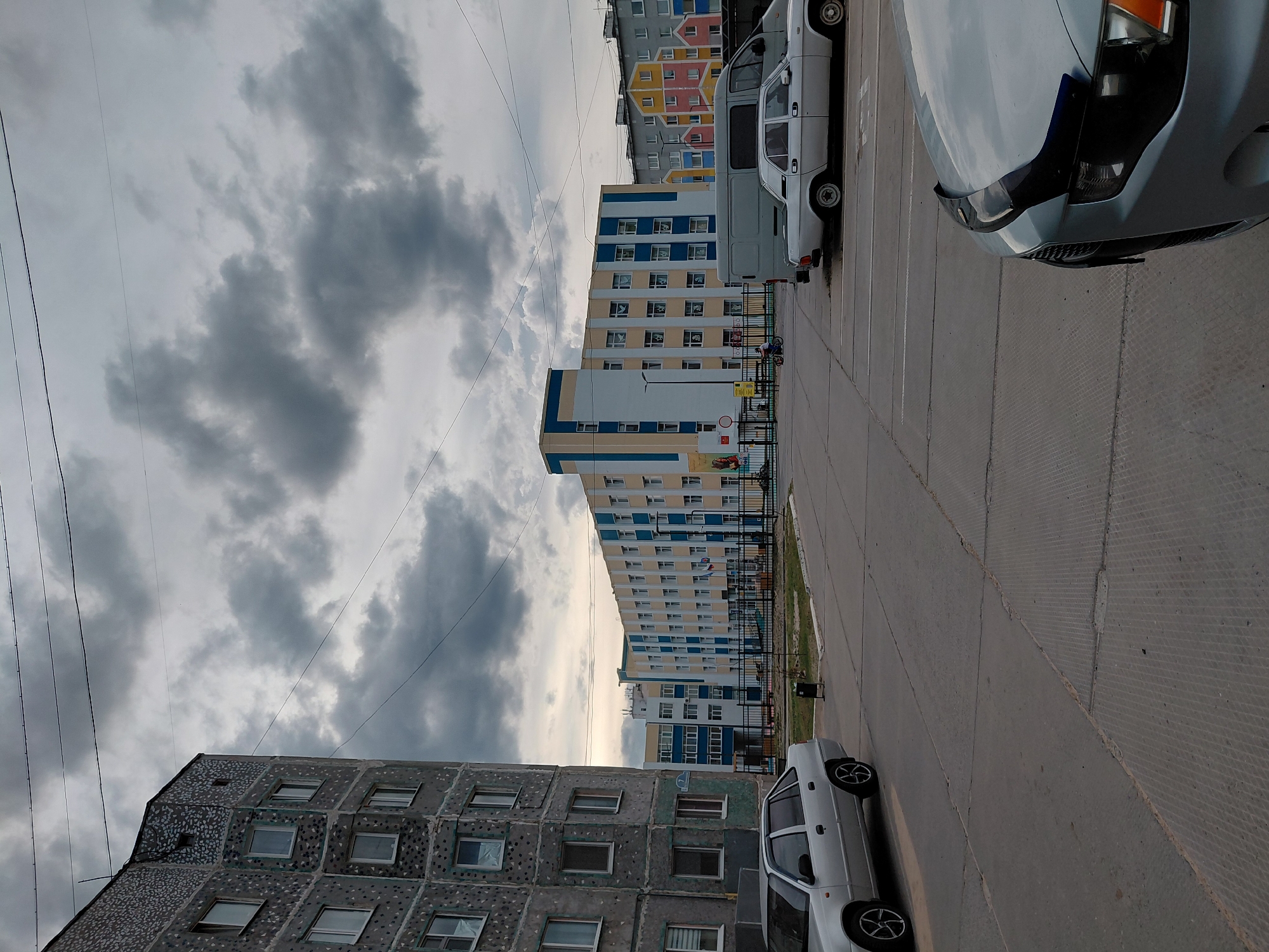 This photograph captures an urban residential scene in Nadym, Russia, under an overcast sky. The image is dominated by multi-story buildings and parked vehicles on a concrete ground.
In the foreground to the left, a grey residential building with a distinctive textured or patterned facade and numerous windows is prominent. To its right, closer to the center, a white compact car is parked on the concrete pavement.
Further into the mid-ground and background, a series of multi-story apartment buildings line what appears to be a street or large parking area. These buildings showcase varied architectural styles and color palettes; some are painted in blue and yellow accents, while others further back feature a mix of yellow, pink, and grey. A small, indistinct flag (possibly Russian) is visible on a balcony of one of the colorful buildings.
On the right side of the frame, several vehicles are parked on the concrete. The front section of a light-colored car (possibly silver or light blue) is seen in the immediate foreground, and a white van is parked behind it. The ground consists of large, textured concrete slabs, indicative of a street or parking lot.
The sky above is heavily overcast with dense, dark grey clouds, suggesting a cloudy daytime. Multiple thin power lines traverse the sky, especially noticeable in the upper left quadrant. No people are distinctly visible, but a small child's scooter or bicycle is present near a fence in the mid-ground, implying a residential environment. The scene depicts a typical, quiet day in a Russian city. No legible text is clearly visible on any signs or objects.