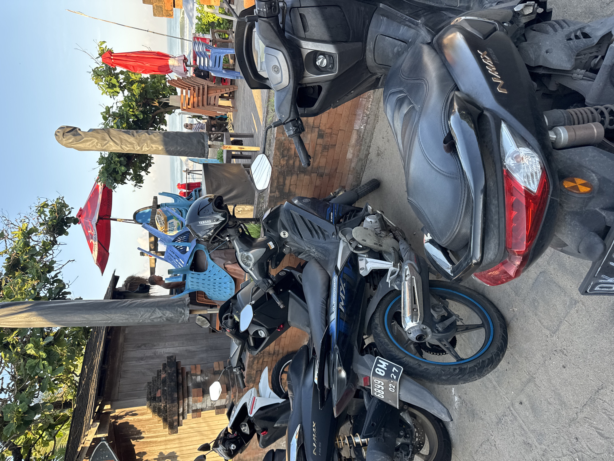This image captures a vibrant street scene in Kuta, Indonesia, characterized by parked motorcycles and outdoor eateries near a coastline. The time of day appears to be late morning or afternoon, with bright sunlight creating clear skies and casting distinct shadows.
In the foreground, several scooters are parked, dominating the lower and right portions of the frame. A prominent black Yamaha NMAX scooter is seen, with "NMAX" branding visible on its side. Another Yamaha scooter, possibly a Jupiter MX based on visible text, is parked alongside, featuring a license plate that reads "6899 OM 02.27." A black helmet with "YAMAHA" written on it rests on one of the bikes. A bottle of "AQUA" drinking water is visible on a seat.
Beyond the parked vehicles, the scene opens up to a row of establishments with outdoor seating. Red sun umbrellas provide shade over blue plastic chairs and wooden tables. The ocean is visible in the background, confirming a beachside location. Scattered individuals can be seen in the background, including one person wearing a striped shirt seated at a table. A tall, grey cylindrical object, possibly a covered flagpole or parasol, stands upright. A traditional Balinese-style building with a wooden facade and intricate brickwork is also visible, adding to the local atmosphere. The overall impression is one of a lively, casual coastal area.