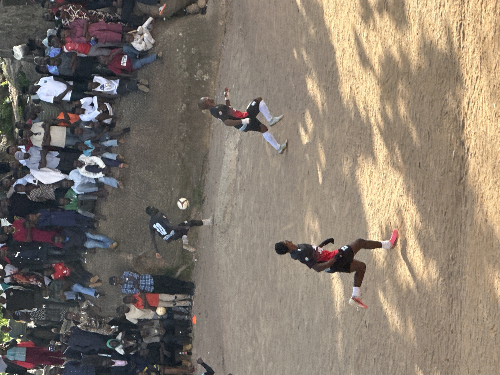The image depicts a dusty outdoor football pitch, possibly in the city of Jos, Nigeria, where a crowd has gathered to watch a match. Several individuals are actively engaged in the game, while a larger group of spectators are positioned on a rocky incline overlooking the playing area. The spectators are dressed in a variety of casual attire, and the overall atmosphere appears to be relaxed and communal. Sunlight casts long shadows across the field, suggesting it is either early morning or late afternoon. The terrain is rough and uneven, characteristic of many informal sporting venues. The presence of a crowd and the game in progress indicate a local event or gathering.