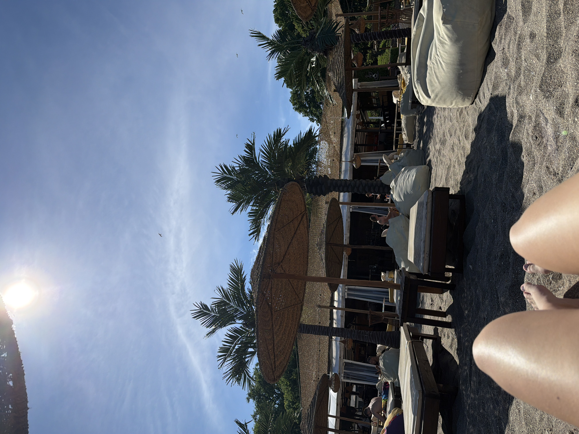 The image depicts a sunny beach scene at a resort in Burgas, Bulgaria. In the foreground, the legs and feet of a person are visible, suggesting they are lying on the sand. The middle ground features several sun loungers and large, straw-covered umbrellas, providing shade. Palm trees are scattered throughout the scene, adding to the tropical atmosphere. In the background, a structure that appears to be a restaurant or bar is visible, with patrons seated at tables. The sky is a clear blue with a few wispy clouds and the sun shining brightly. Several birds are also visible flying in the sky. The overall impression is one of a relaxed and enjoyable beach environment.