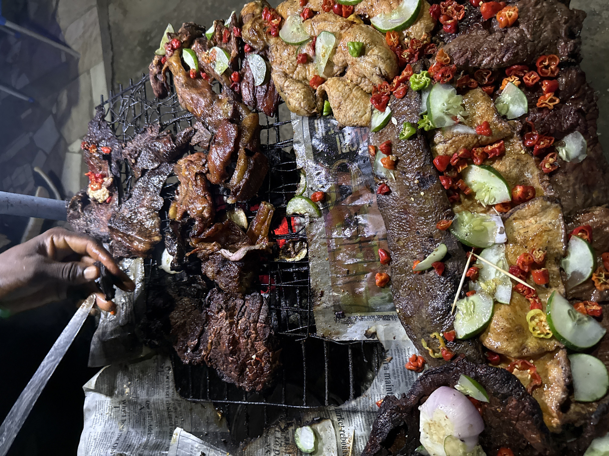 In Abuja, Nigeria, a cook is preparing a variety of grilled meats, including what appears to be beef and chicken, on a metal grate over hot coals. The meats are seasoned and garnished with sliced cucumbers and red chilies. Some of the meat is being sliced by the cook with a knife. The scene appears to be outdoors at night, with newspapers used as a base beneath the grilling setup.