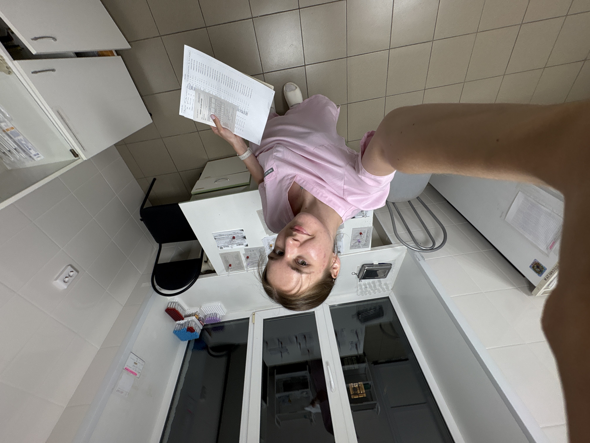 A person in a pink scrub suit is holding a document and standing in a room with white tile floors and white walls. The room appears to be a medical or laboratory setting, with cabinets, a sink, and laboratory equipment visible. The person is taking a selfie and is partially blocking the right side of the image with their arm. Outside a window, there is a reflection of a building, suggesting an urban environment. No location details, specifically relating to Perm, Russia, can be extracted from the image.