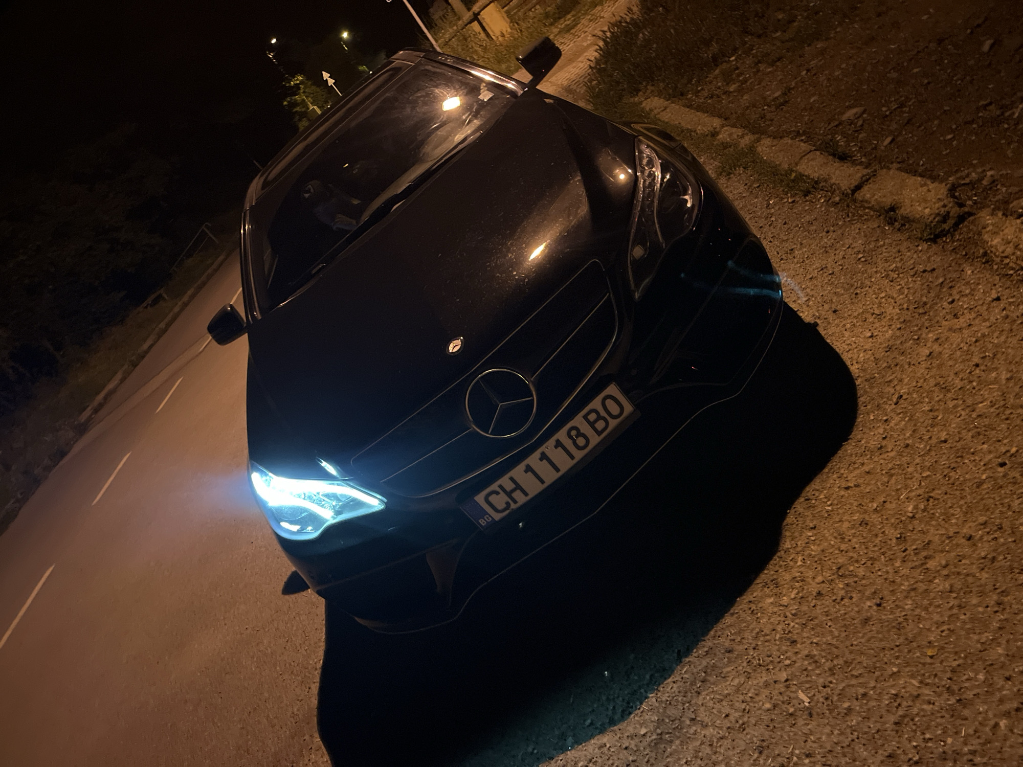 The image captures a dark-colored Mercedes-Benz car parked on the side of a road at night. The car is positioned diagonally, with its front facing towards the bottom left of the frame. Its bright headlights are illuminated, casting light onto the asphalt road surface directly in front.
The scene is dark, indicating nighttime, with streetlights visible in the distance providing additional illumination to the surroundings. On the right side of the car, there is a rough, gravelly area bordering a curb, suggesting a roadside or unpaved shoulder. On the left, a paved road stretches into the distance with faint white lane markings visible. Trees and foliage are present in the dark background, along with distant light sources, possibly other streetlights.
Notable details include the prominent Mercedes-Benz logo on the car's front grille. A European-style license plate is clearly visible, displaying the country code "BG" for Bulgaria and the alphanumeric sequence "CH 1118 BO." The car's paintwork appears dark, possibly black, with some dust or reflections visible on the hood. A white arrow sign on a pole is faintly discernible in the upper background, pointing to the right. The overall impression is a static, nighttime street scene centered around the parked car.