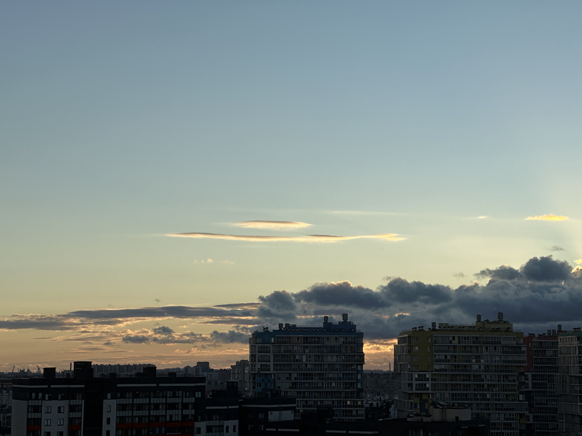 The image depicts a cityscape at sunset. In the foreground, several multi-story residential buildings are visible, some with dark facades and others with lighter, colored sections. The buildings are densely packed, suggesting an urban environment. Above the buildings, a sky with various cloud formations is present. Some clouds are dark and dramatic, while others are wispy and illuminated by the setting sun, creating a warm glow. Long, thin lenticular clouds are particularly noticeable in the upper portion of the sky. The overall lighting suggests either early morning or late evening. The location is identified as Novosergievka, Russia. There are no people or visible actions taking place in the image; it is a static view of the urban landscape and sky.