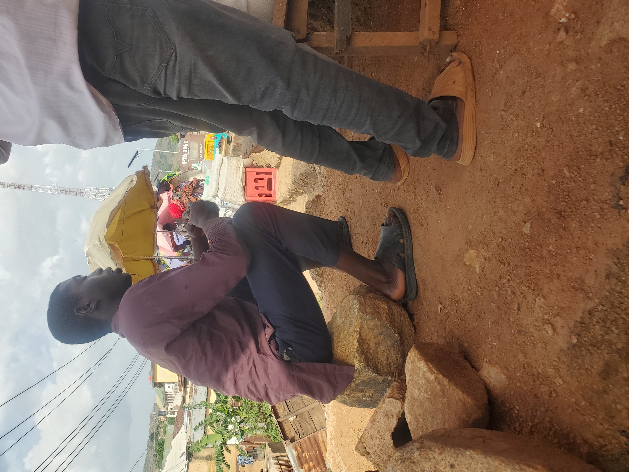 The image, taken in Gwong, Nigeria, depicts a scene on a dirt ground, likely outdoors given the daylight and the presence of a utility pole in the background. Two individuals are partially visible. In the foreground, a person wearing a maroon long-sleeved shirt and dark pants sits on large rocks. This individual is looking upwards and to the left, with a neutral expression. Their feet are clad in black sandals.
Behind this seated person, another individual's legs are visible. This person wears dark grey jeans and light brown sandals. Their stance suggests they are standing.
In the mid-ground, a yellow umbrella provides shade over a group of people and what appears to be market stalls. Bags and crates are visible, suggesting a marketplace setting. A red crate is placed on the ground near the standing person's legs. In the distance, a transmission tower stands tall against a bright, cloudy sky. The overall atmosphere suggests a busy outdoor environment, possibly during the daytime. The terrain is dry and dusty, consistent with an arid or semi-arid climate.
