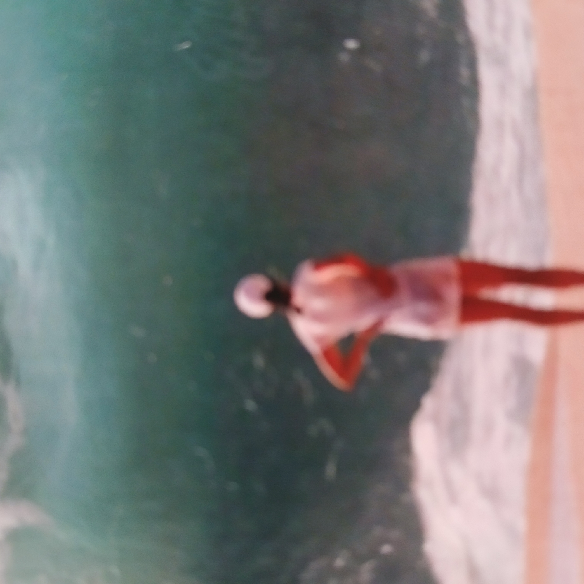 A person wearing a light-colored cap and a matching dress stands on a sandy beach, facing away from the camera towards a large wave. The water is a deep teal color and appears to be breaking. The person's hands are on their hips. The sky is not visible. The scene is extremely blurry and lacks detail, making it impossible to ascertain the specific location within the city of Samarsky, Russia, or any further identifying features of the person or their surroundings.