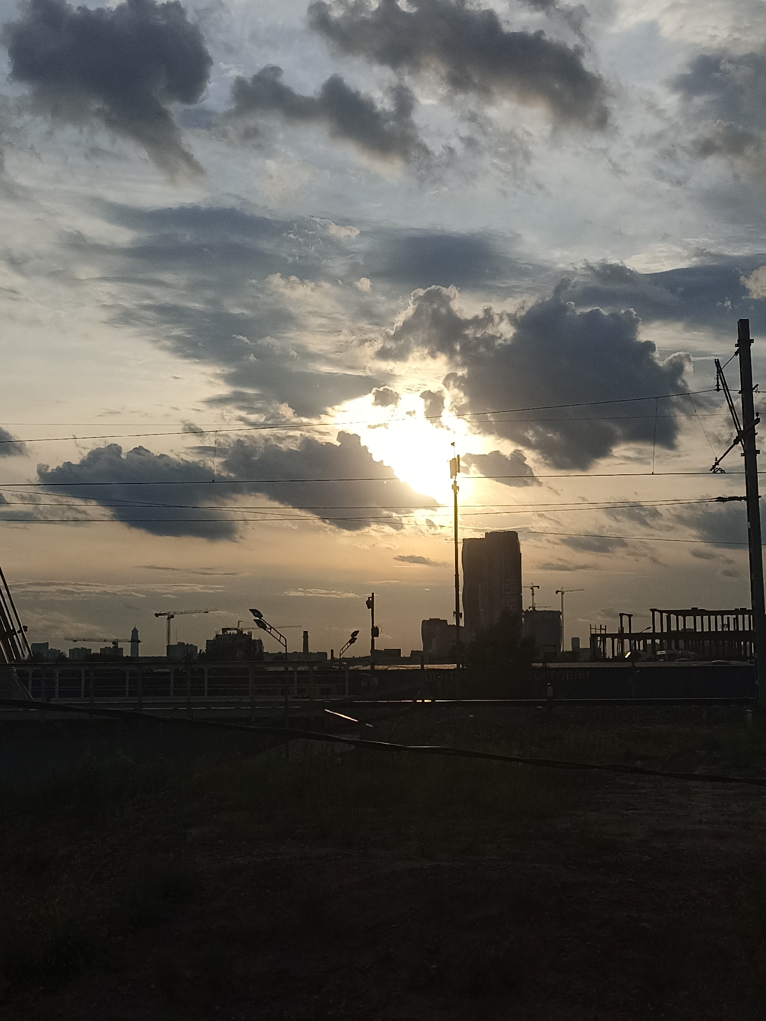 A wide shot captures a cloudy sky with the sun partially obscured by dark clouds, casting a warm, golden light. In the mid-ground, a city skyline is visible, with several tall buildings and construction cranes indicating ongoing development. Power lines run across the frame, supported by utility poles. The foreground is dominated by dark silhouettes of vegetation and possibly a railway track. The scene appears to be at dusk or dawn, with the low sun creating dramatic lighting.