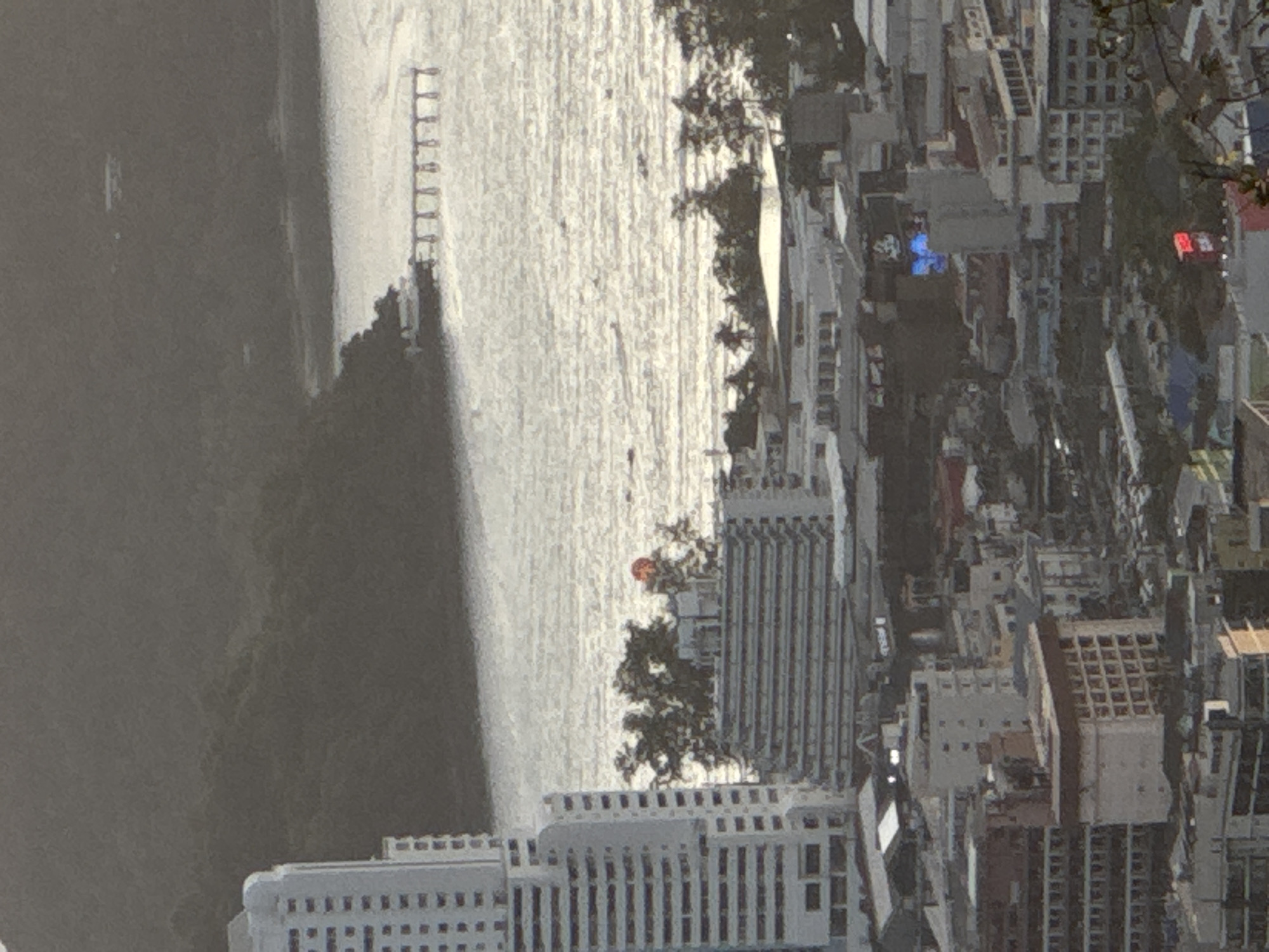 This is a daytime, aerial, long-shot photograph of a coastal city, likely Patong, Thailand, featuring a densely packed urban landscape with numerous buildings of varying heights. In the background, a dark, tree-covered hillside slopes down towards a body of water that reflects the sunlight. A long, thin pier extends out over the water from the shore.
The foreground is dominated by the city. A very tall, white, multi-story building with a grid-like facade is prominent on the left. To its right, a mid-rise building with a similar facade is visible. Further into the city, a cluster of lower-rise buildings and structures are densely packed. Some buildings appear to be hotels or apartment complexes with repetitive window patterns. There are also some roads or parking areas visible with parked cars.
In the mid-ground, several buildings stand out. One has a distinctive stepped or terraced design. Another building has a large, illuminated sign with blue lettering. A smaller building below this has a red illuminated sign. A bright red, circular object, possibly a sign or light, is visible atop some trees in the middle distance.
The water appears slightly choppy, with visible ripples and streaks from the sunlight. A few small boats are scattered across the water. The overall lighting suggests it is either late afternoon or early morning, with the sun casting a bright glare on the water. There are no visible people in the frame. The scene depicts a developed coastal urban area with a significant presence of multi-story buildings and a connection to the sea.