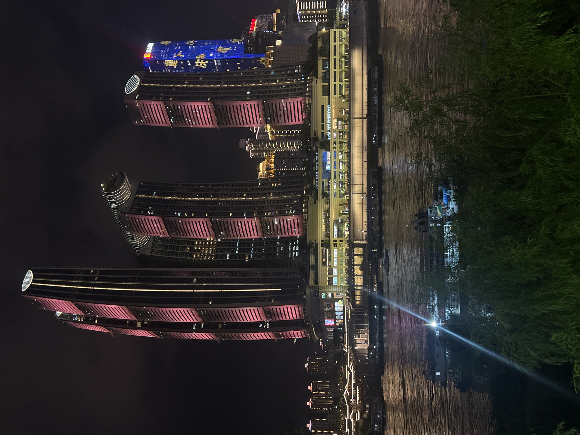 This image depicts a nighttime cityscape along a waterfront, likely in Jiangbei District, Chongqing, China, given the prominent architecture which is characteristic of the area, particularly the "Lotus Hill" skyscrapers. Several tall, modern buildings are illuminated with vibrant pink and blue lights. The central focus is a cluster of skyscrapers, with some featuring distinctive curved designs and patterned lighting. A lower, horizontally oriented building complex runs along the waterfront, also brightly lit. In the foreground, a wide river or bay reflects the city lights, with several boats of various sizes visible on the water. A dense canopy of green foliage, likely trees or large shrubs, occupies the bottom portion of the image, creating a natural frame and contrasting with the urban structures. The overall impression is one of a bustling, illuminated urban environment at night.