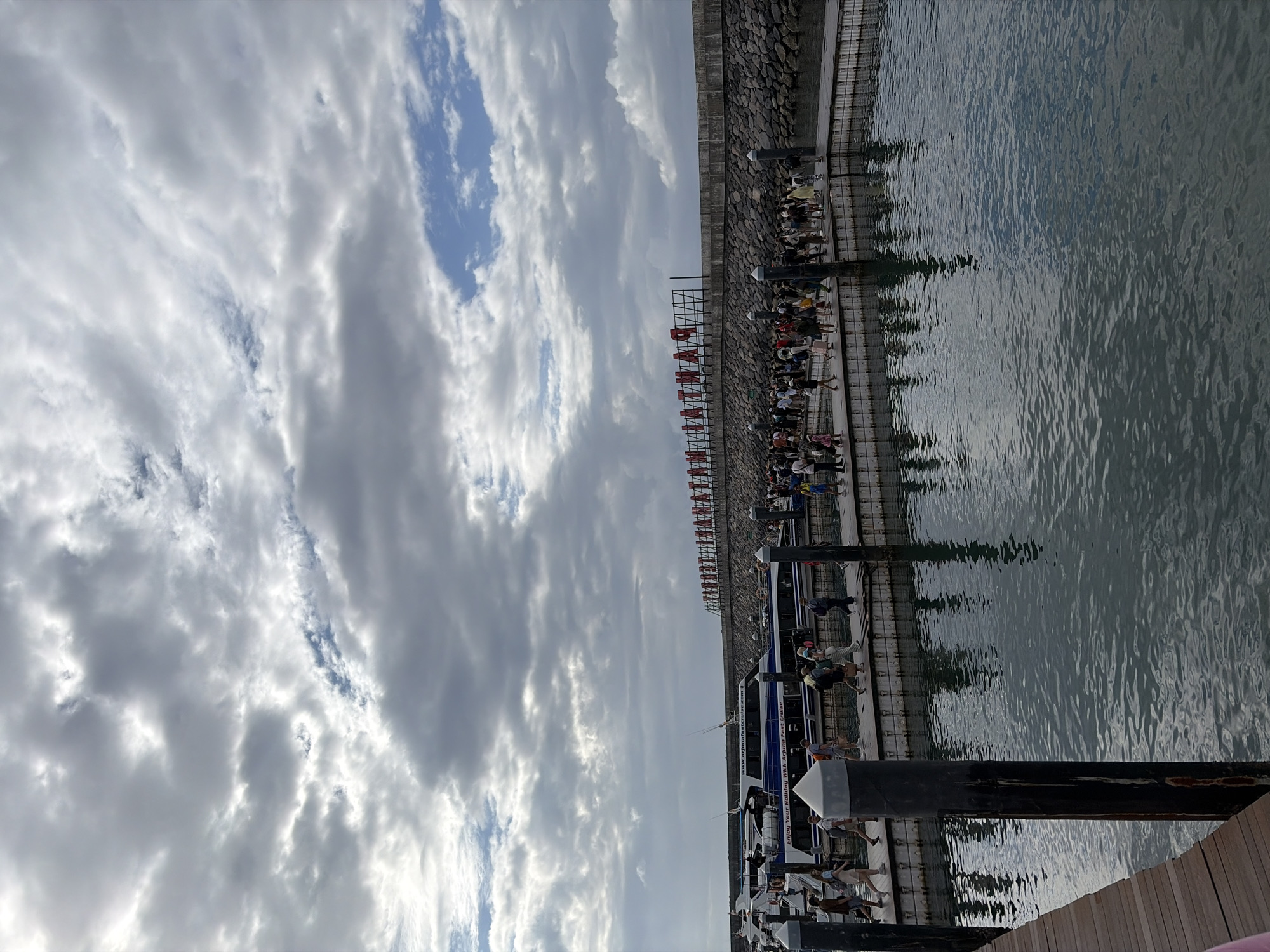 The image depicts a bustling harbor scene, likely in Denpasar, Indonesia, with a focus on a pier filled with people and a fast boat. The sky above is overcast with dramatic clouds, casting a diffused light over the scene. In the background, a large sign with red lettering spells out "SANTIKA" horizontally. A concrete wall lined with rocks forms the harbor's edge, separating the water from the land beyond.
The pier itself is constructed of wood and lined with dark posts. A group of people, casually dressed and appearing to be tourists, are walking along the pier. They are diverse in age and gender, carrying bags and backpacks. Their activity suggests they are either disembarking from or preparing to board one of the boats docked at the pier.
A prominent fast boat, labeled "Enjoy Your Holiday With Arjuna Fast Cruise," is docked alongside the pier. The boat is white and blue and appears to be modern and well-maintained. Other boats are visible docked behind it, suggesting a busy ferry terminal or harbor.
The water in the foreground is dark and reflects the cloudy sky and the surrounding structures, creating a somewhat moody atmosphere. The overall scene conveys a sense of travel and activity at a popular tourist destination.