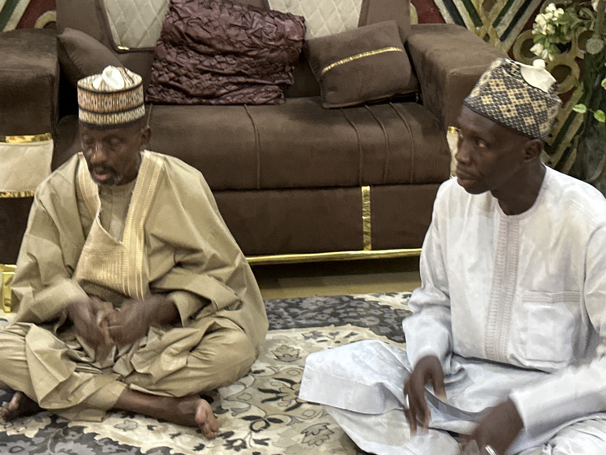 The image depicts two elderly men sitting cross-legged on an ornate rug indoors. The man on the left is wearing a tan robe and a cylindrical hat with a light brown and dark brown band. He is barefoot. The man on the right wears a white robe and a similar hat with a checkerboard pattern. Behind them is a brown sofa with gold accents, adorned with brown and quilted beige pillows. To the right, a vase holds white flowers against a backdrop of geometric wall decor. The location is likely a residential setting, city and state Unknown.