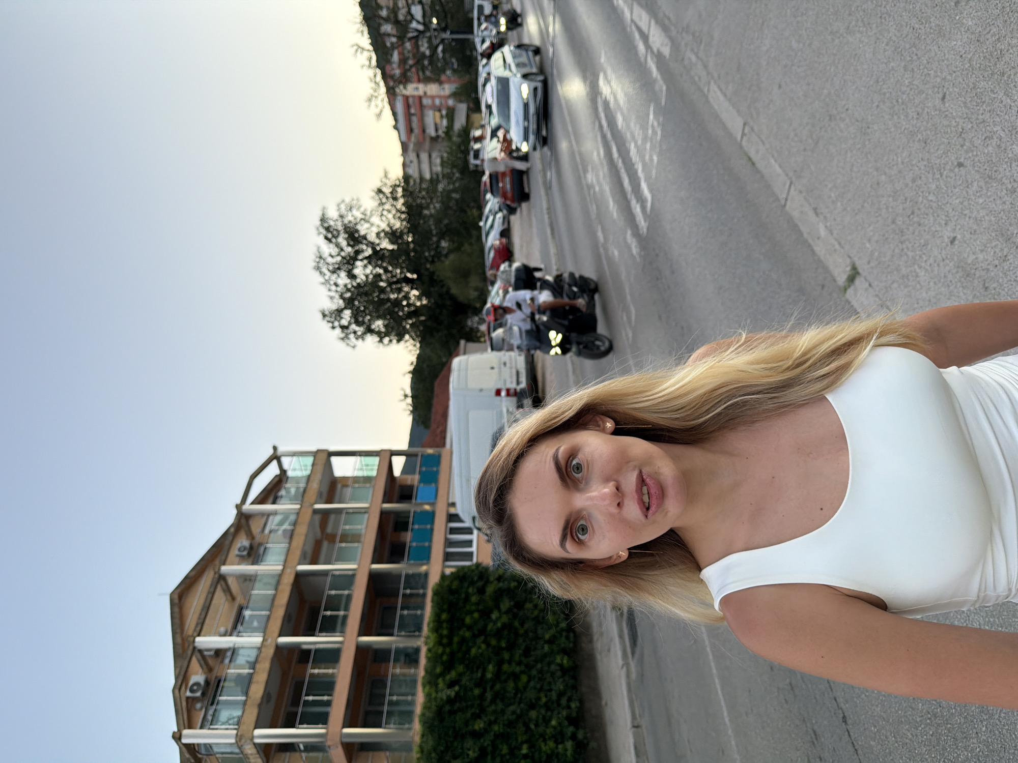 A blonde woman in a white tank top stands on a street in Budva, Montenegro. She has long blonde hair and large, surprised eyes. Behind her, a multi-story building with balconies and air conditioning units is visible. A white van is parked in front of the building. The street is lined with cars, and a scooter is being ridden by a person wearing a red helmet. Trees and other buildings can be seen in the background under a clear sky. The image appears to have been taken at dusk.