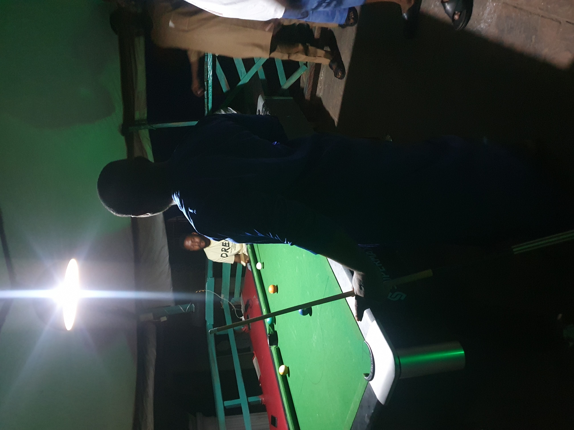 In Lokoja, Nigeria, a group of men are gathered around a green billiard table illuminated by a bright overhead light. One man, wearing a blue shirt, stands with his back to the camera, holding a billiard cue. Another man, in a light-colored t-shirt with "DREAM" printed on it, is visible further back, looking towards the table. A third man, in a tan outfit, is partially visible to the right. Several billiard balls are scattered across the table, with one blue ball in the foreground. The setting appears to be an outdoor or semi-outdoor area, with a turquoise railing in the background. The atmosphere is casual, with the focus on the game of billiards.