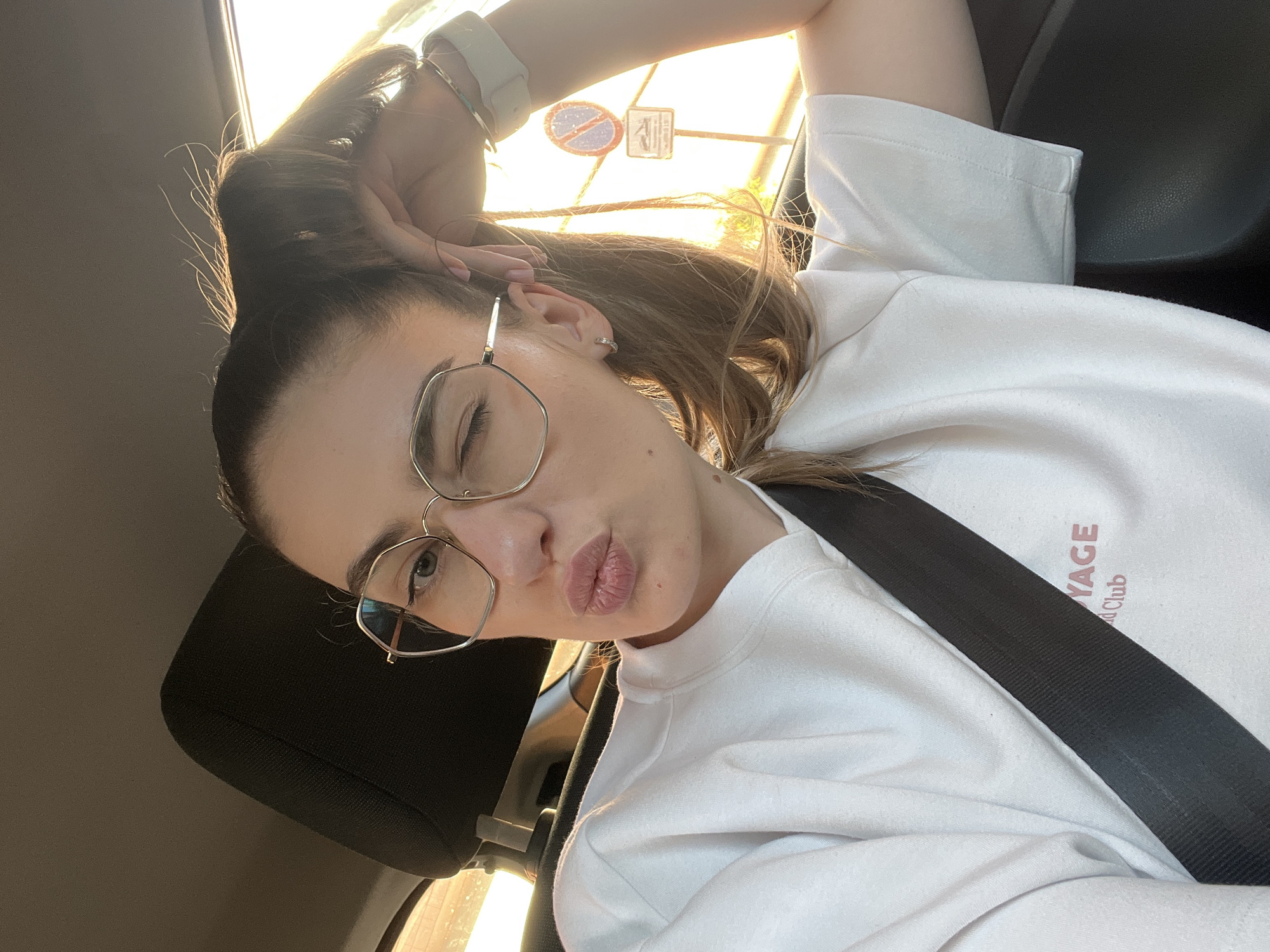 A woman with light brown hair pulled into a high ponytail is in the driver's seat of a car. She wears gold wire-rimmed glasses and a white t-shirt with a seatbelt across her chest. She is winking and pursing her lips. The window is behind and above her with sunlight streaming in and reflecting off a road sign outside. The city is Unknown, Unknown.