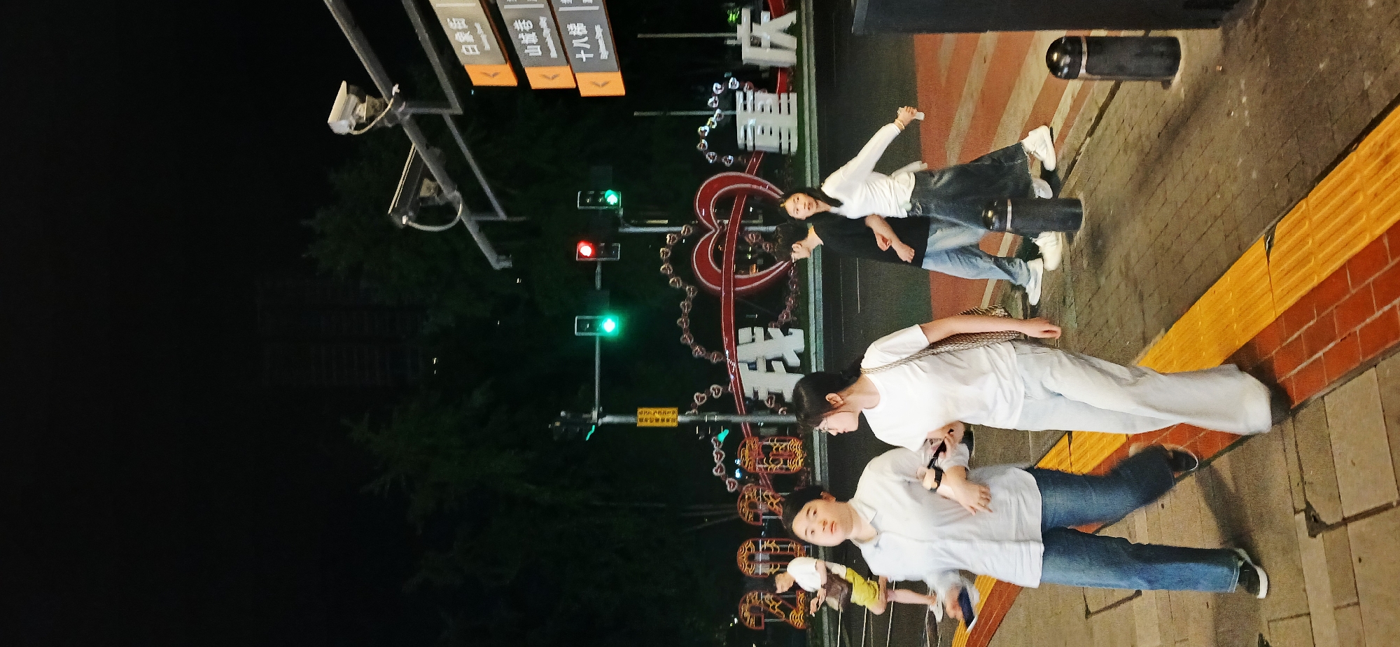 People stand on a brick-paved sidewalk at night in Yuzhong District, China. A street sign overhead lists local streets, including "Shiba Ti." A heart-shaped neon sign decorated with strings of lights is visible in the background, along with traffic lights. Three women are in the foreground, and one man stands behind them. All face forward.