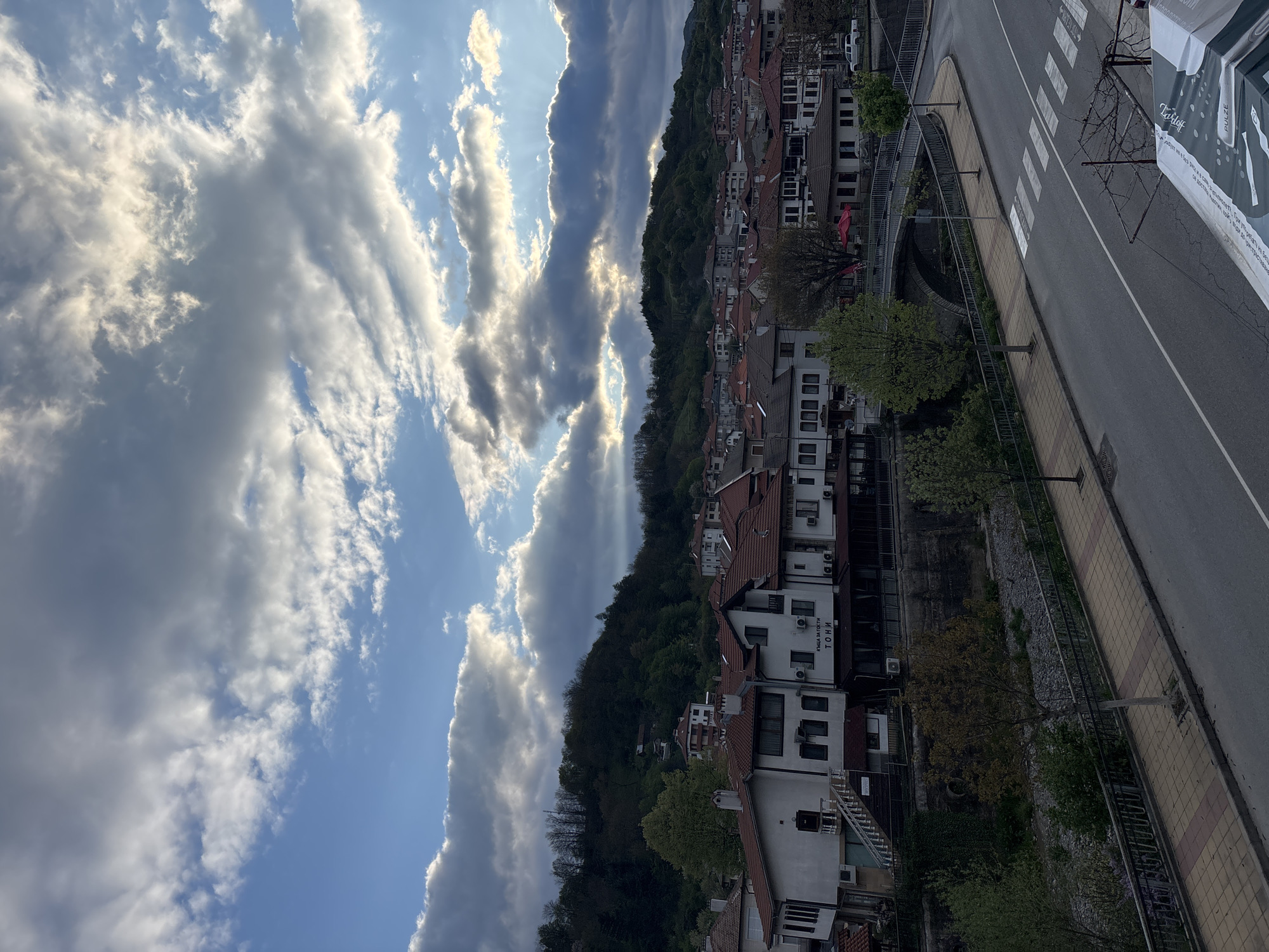 The image shows a vertical view of Zlatograd, Bulgaria. The sky is filled with white clouds and sunbeams. A tree-covered hill lies between the sky and the town below. The town is made up of buildings with red roofs, interspersed with greenery. A road runs parallel to the buildings, with a crosswalk visible. A sidewalk separates the road from the buildings.