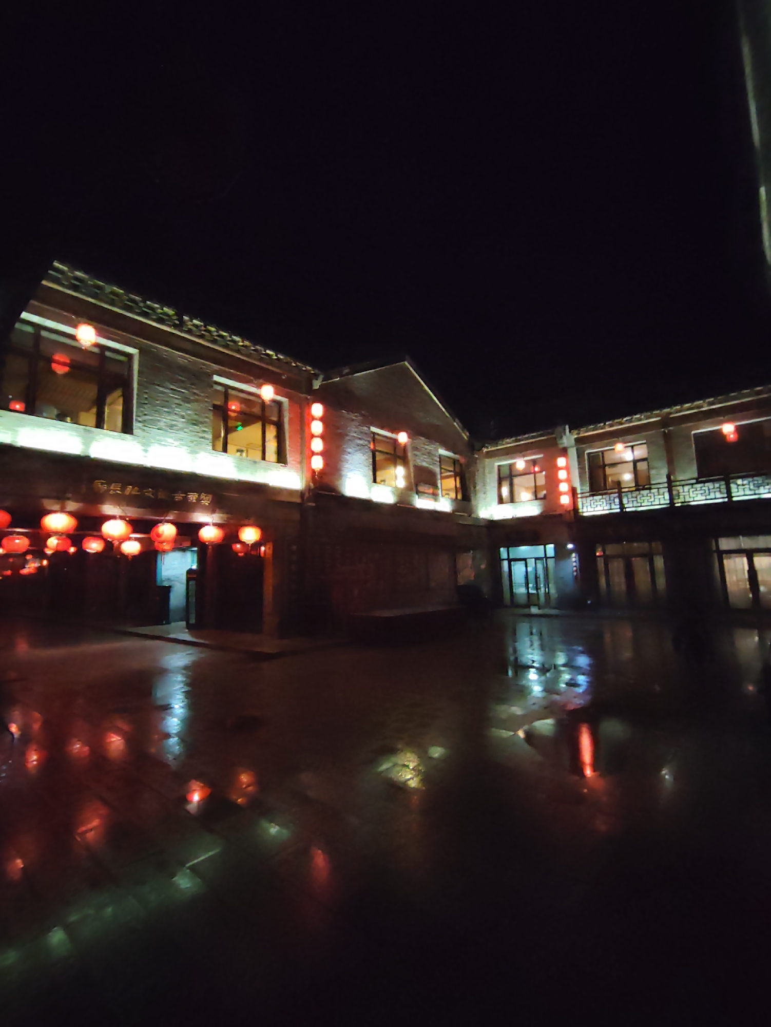 The image depicts a courtyard at night, illuminated by red lanterns and interior lights from a building complex. The ground appears wet, reflecting the lights. The architecture suggests a traditional Chinese style, with multi-story buildings and decorative elements. A sign with Chinese characters is visible above an entrance. The scene is set in Lindian, China, likely a commercial or hospitality area given the inviting lights and building design. There are no people or animals visible in the frame. The overall atmosphere is serene and festive.