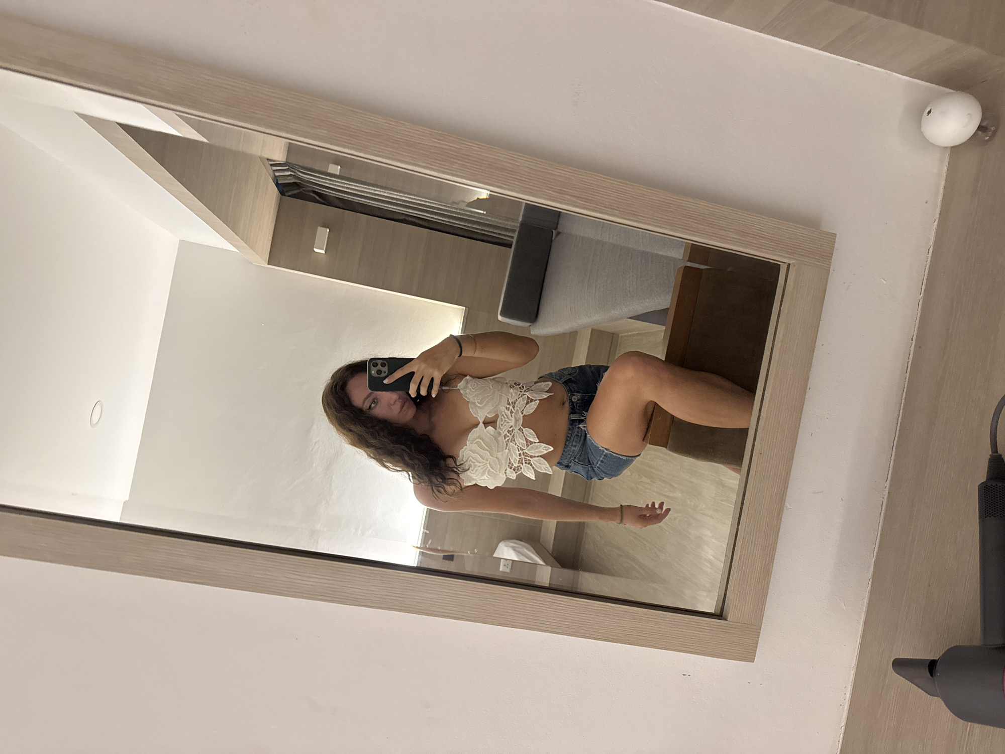 In a room with white walls and light wood-paneled accents, a woman with long, dark, wavy hair is taking a mirror selfie. She is wearing a white, floral-patterned lace bralette and distressed denim shorts. Her left leg is crossed over her right, and she is holding a black smartphone in her right hand to capture the reflection. Behind her, a mirror reflects a bed with white linens, a bedside lamp, and a gray upholstered headboard. The room appears to be a hotel room. A hairdryer is visible in the foreground of the image. The scene suggests the woman is staying in a hotel in Ko Samui District, Thailand.