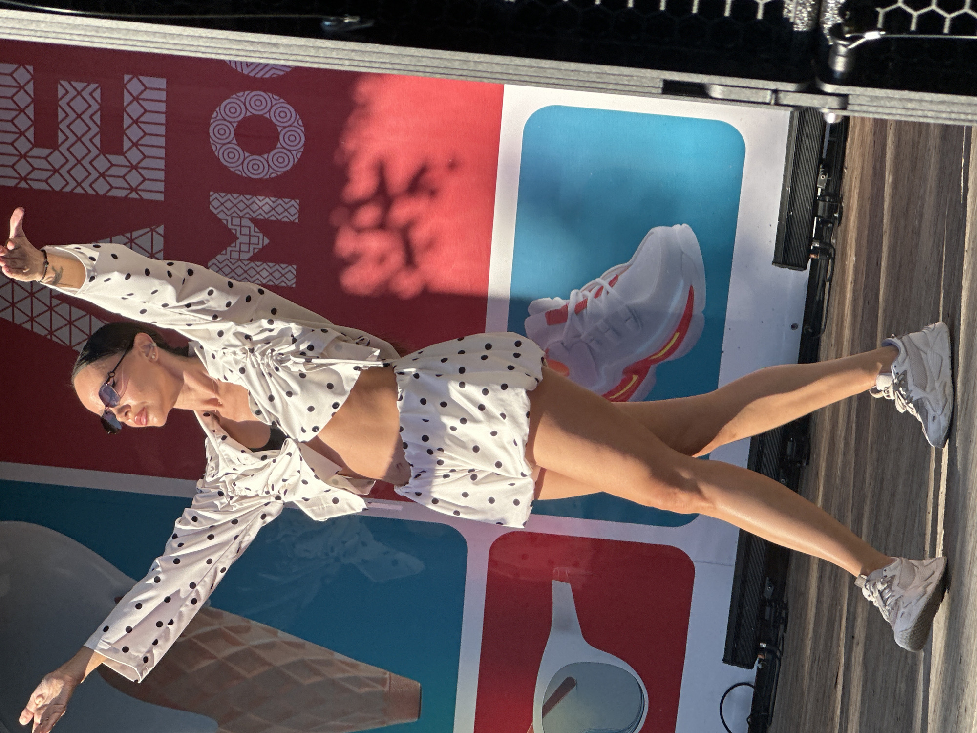 The image depicts a woman in a white and black polka dot outfit, consisting of a cropped top and shorts, posing with her arms outstretched. She is wearing sunglasses and sneakers. The background features a large banner with colorful graphics and text, including the word "MOSCOW" in stylized lettering. The scene appears to be an outdoor event or performance, possibly in a public square or park in Moscow, Russia, given the prominent display of the city's name. The lighting suggests it is daytime, with sunlight casting shadows.
