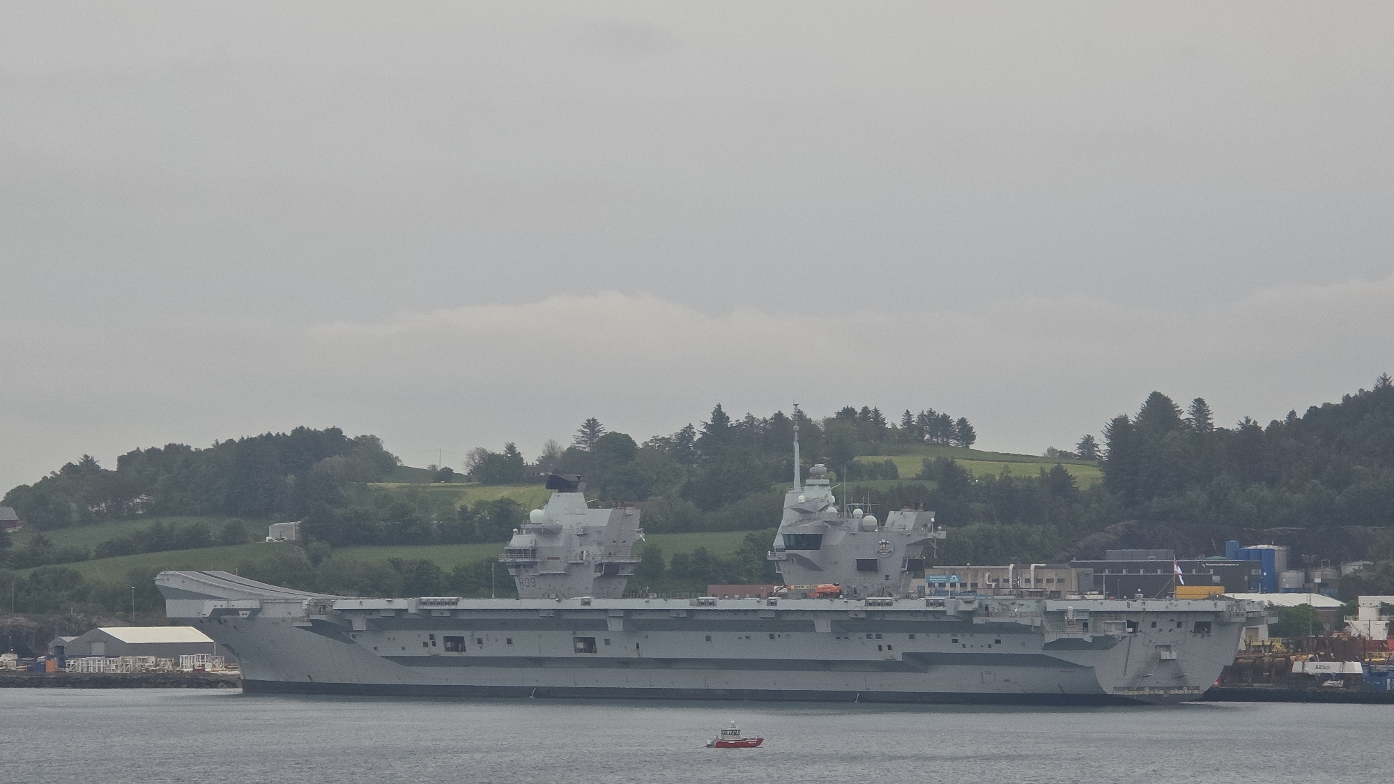 The image displays a large grey aircraft carrier, identified as a Queen Elizabeth-class warship, with its distinctive twin island superstructure and a ski-jump ramp at the bow. The pennant number "R08" is clearly visible on the port side of the forward island, indicating the vessel is HMS Prince of Wales. A British White Ensign flag flies from the stern of the ship.
The aircraft carrier is situated in a body of water, likely a fjord or bay, with its starboard side facing the viewer. In the mid-ground, a small red boat is visible on the water near the larger vessel. The background consists of verdant, undulating hills densely covered with coniferous trees, stretching along the coastline. Some scattered buildings and infrastructure are present on the shore. The sky is uniformly overcast with grey clouds, creating a diffused light over the scene. The water appears calm. The photograph captures the immense scale of the warship against the natural, hilly landscape.