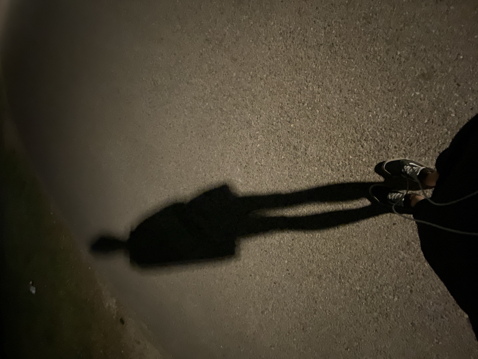 A shadow cast by a person standing on a paved surface is visible in this low-light photograph. The person is wearing shoes and dark clothing. The surface is rough and appears to be asphalt or concrete. The setting appears to be outdoors, possibly a street or walkway, at night.