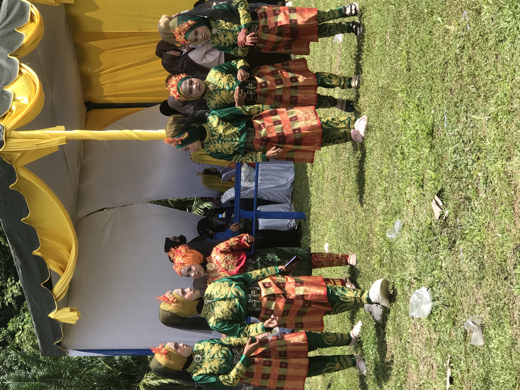 Six young girls, dressed in traditional green and gold embroidered tops and skirts, stand on a grassy field. They are all wearing headscarves, some adorned with orange floral decorations. The girl on the far left and the two girls on the right are wearing green headscarves, while the middle girl is wearing a yellow headscarf. The girl second from the right wears an orange headscarf. The girls are posing for a photo. In the background, a tent with yellow and white fabric is set up, likely for an event. Trees are visible behind the tent, suggesting an outdoor location. The ground is covered in green grass with some scattered litter, including plastic bags and cups. The context of the image suggests a cultural event or performance in Cotseutui, Indonesia.