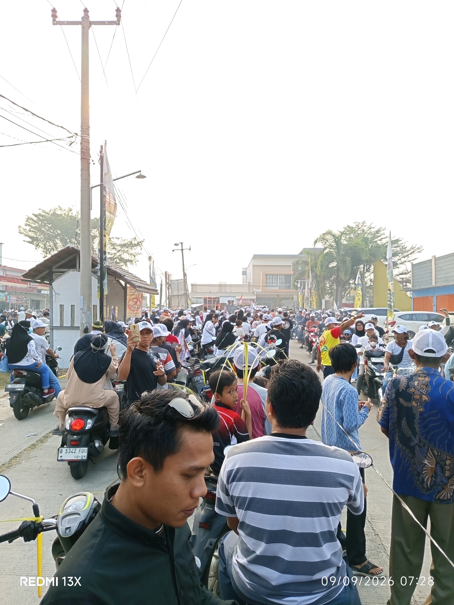 The image captures a street scene in Karanganyar, Indonesia, likely during an event given the large gathering of people on motorcycles. The sky is bright and overcast, suggesting it is daytime. In the foreground, a man in a black jacket is seen from the side, looking down. Behind him, other individuals are on motorcycles, some with passengers.
Further down the street, a crowd of people, many wearing white hats or head coverings, are visible. They are predominantly on motorcycles, creating a procession or gathering. Some individuals are holding palm fronds, which might indicate a religious or cultural event. Flags and banners are also present along the street, some displaying text and images, hinting at a public event or rally.
The environment includes buildings on either side of the road, a utility pole with electrical wires, and street lamps. The road surface appears to be asphalt. The license plates of some motorcycles are visible, with one reading "B 5342 FUU" and another "3335 F." The time stamp in the lower right corner indicates "09/09/2026 07:28." The overall impression is a busy street filled with people participating in a communal activity.