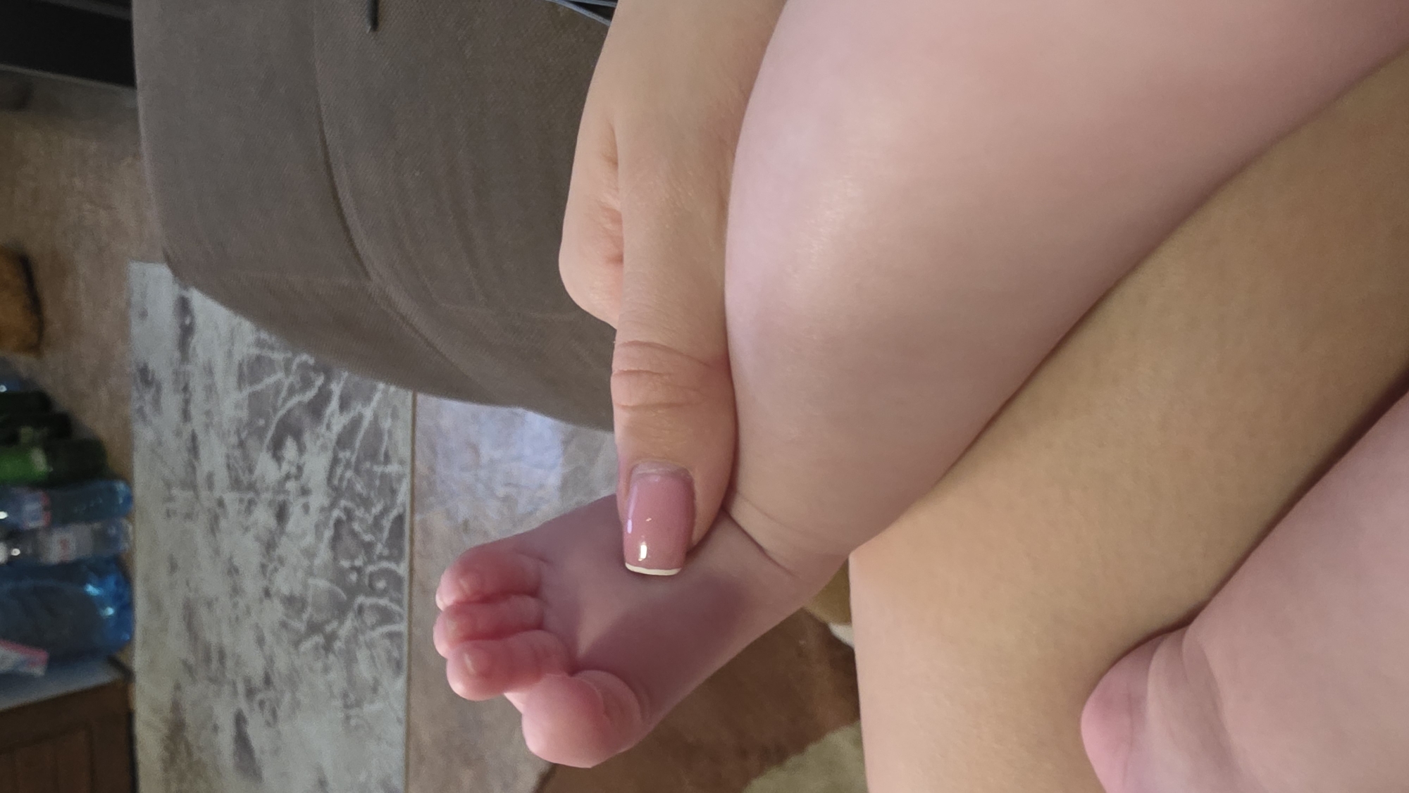 A close-up shot shows a baby's foot being held by an adult's hand. The baby's skin is smooth and pink, while the adult's hand has a manicured nail with a pink base and white tip. In the background, there's a blurry image of a brown couch and a marbled wall or floor. The location is not evident from the image and cannot be determined to be Blagoevgrad, Bulgaria, or any other specific place.