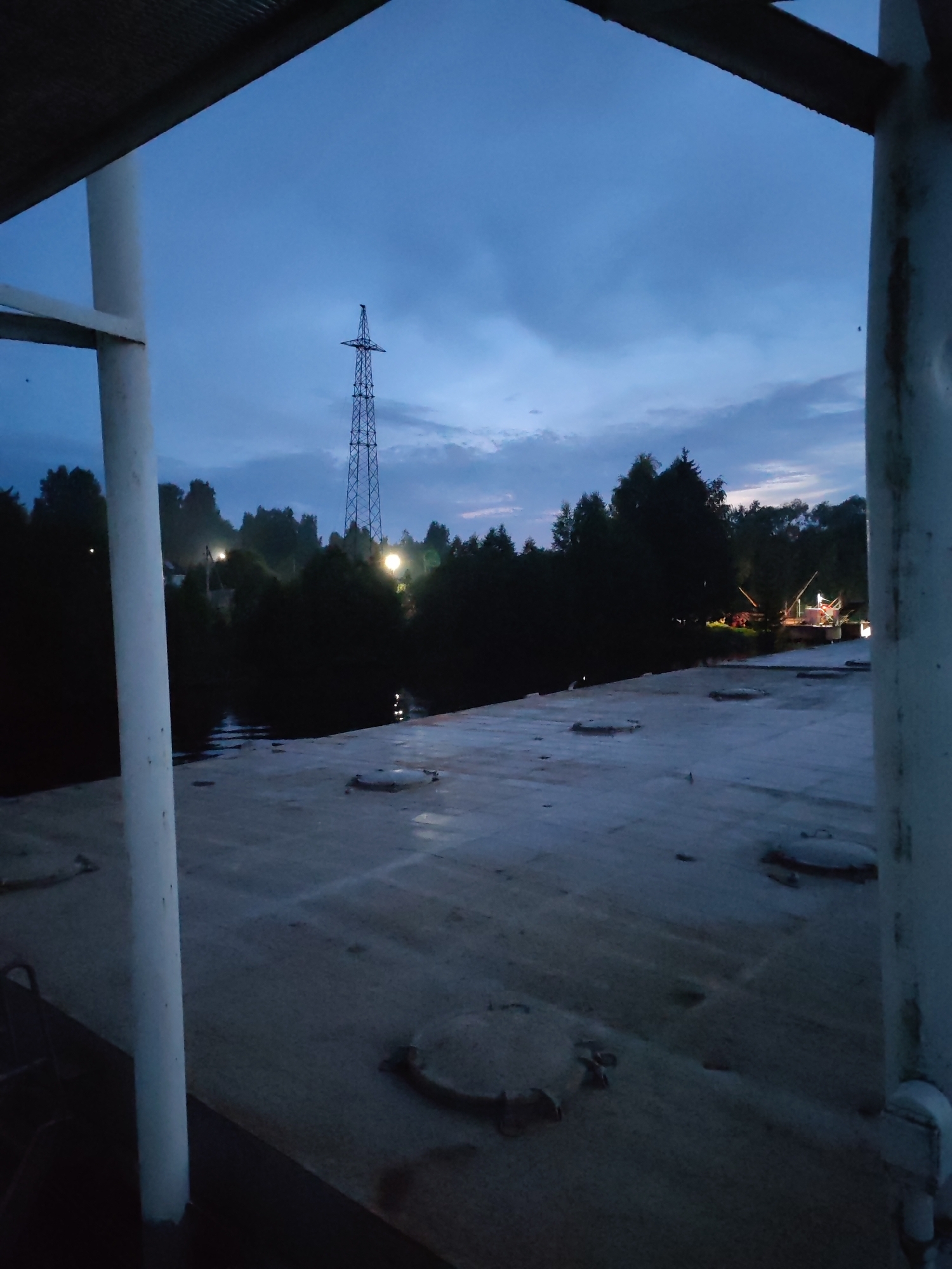 The image depicts a twilight scene over a body of water near Annensky Most, Russia. In the foreground, a light-colored, flat surface, possibly a deck, occupies the lower half of the frame. This surface has several circular, metallic covers or hatches. Two white pillars frame the left and right sides of the image, suggesting a structure or walkway.
Beyond the foreground, a dark body of water is visible, reflecting some lights. The shoreline is lined with dense, dark green trees. In the distance, a tall, lattice-like metal transmission tower stands prominently against the dusky sky. Several artificial lights are visible among the trees, some appearing as bright white or yellow orbs. One cluster of lights on the far right seems to be part of a structure with scaffolding or poles. The sky is a deep blue, transitioning to lighter shades near the horizon, with some faint cloud formations. The overall atmosphere is dim, characteristic of dusk or early evening.