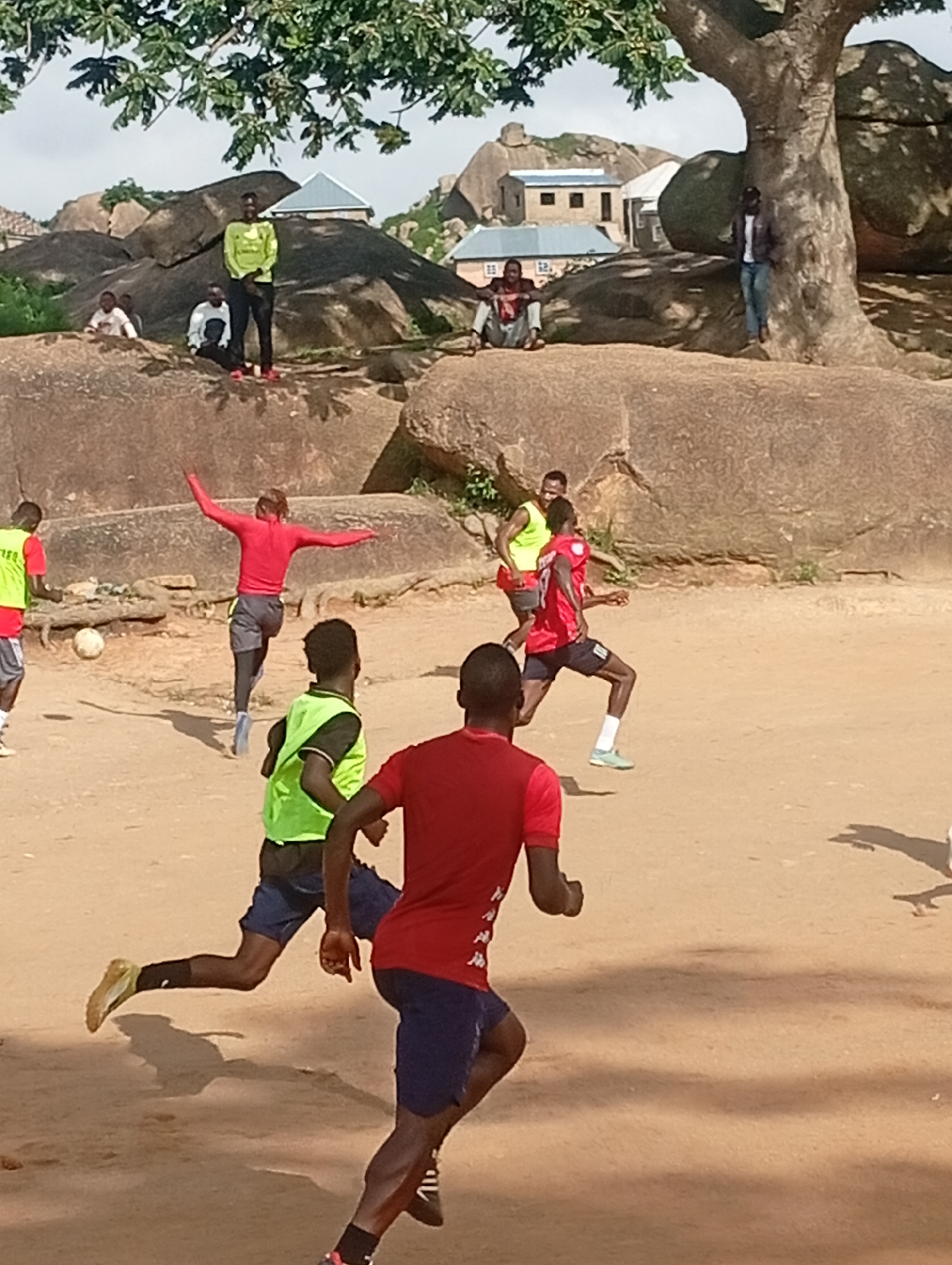 In a dusty, outdoor area with large rock formations and scattered buildings in the background, a group of young men are playing soccer. The foreground shows three men in motion. One man, in a red shirt and blue shorts, is running with his back to the camera, his arms outstretched. To his left, another man in a yellow vest and blue shorts is also running, looking towards the soccer ball. The third man in the foreground, also in a red shirt and blue shorts, is further away and is facing towards the other players. In the background, other men are visible sitting on the rocks and standing near the buildings. The environment appears dry and somewhat arid, with a large tree providing some shade on the left side of the image. The sky is overcast.
