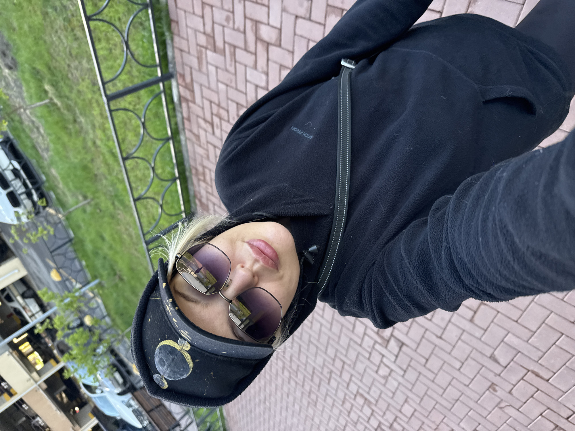 A woman wearing a black fleece jacket, sunglasses, and a black visor with decorations is standing on a paved area. A black metal fence borders a grassy area with trees and parked cars, suggesting an urban setting. The image was taken in Krasnoyarsk, Russia.