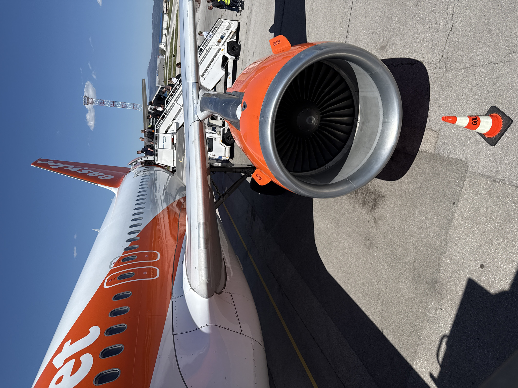 The image captures an EasyJet airplane parked on the tarmac at Sofia Airport, Bulgaria. The aircraft's tail, painted in the airline's distinctive orange and white color scheme, is visible. A jet engine is prominently featured in the foreground, with its silver intake surrounded by an orange nacelle. A set of portable stairs is positioned alongside the aircraft, with passengers and airport personnel visible on the steps. In the background, a control tower and distant hills are visible under a clear blue sky. A small orange and white traffic cone sits on the pavement next to the engine.