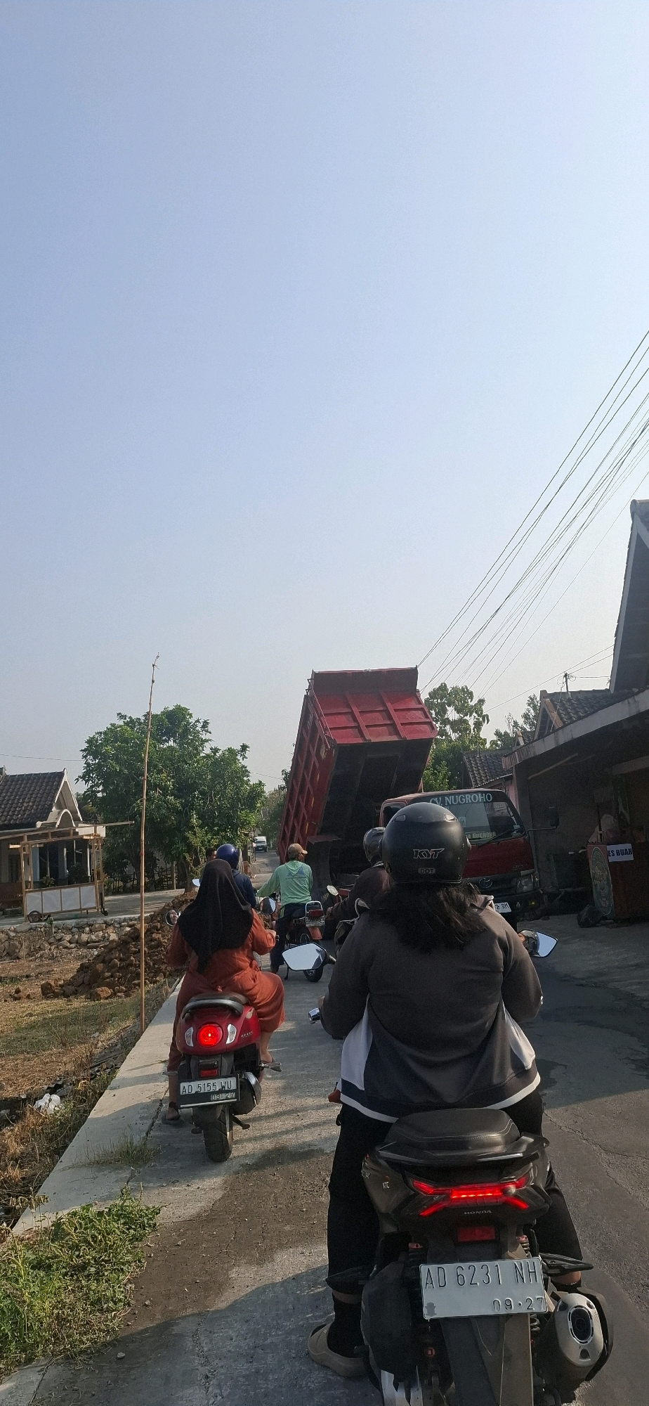 In the city of Desa Sobokerto, Indonesia, a bright red dump truck with its bed raised high is the central focus of this scene, appearing to be in the process of dumping its load. The truck, with "CV. NUGROHO" visible on its side, is positioned on a paved road that curves to the right. Several motorbikes are stopped behind the truck, suggesting a traffic obstruction. On the left side of the road, a pathway borders a construction area with piles of dirt and scattered debris, leading to a small building with a white exterior and a dark roof. Further down the road, more buildings with tiled roofs are visible, interspersed with trees. The sky above is a clear, pale blue. In the foreground, the backs of two individuals on motorbikes are prominently displayed. The person on the right wears a black helmet with "KYT" branding and a dark grey jacket over black pants. The person on the left, wearing an orange headscarf and an orange outfit, is seated on a red scooter. Another individual in a light green shirt on a scooter is visible in the middle distance, also behind the dump truck. The overall atmosphere suggests a typical day in a rural Indonesian setting, momentarily disrupted by the large truck.