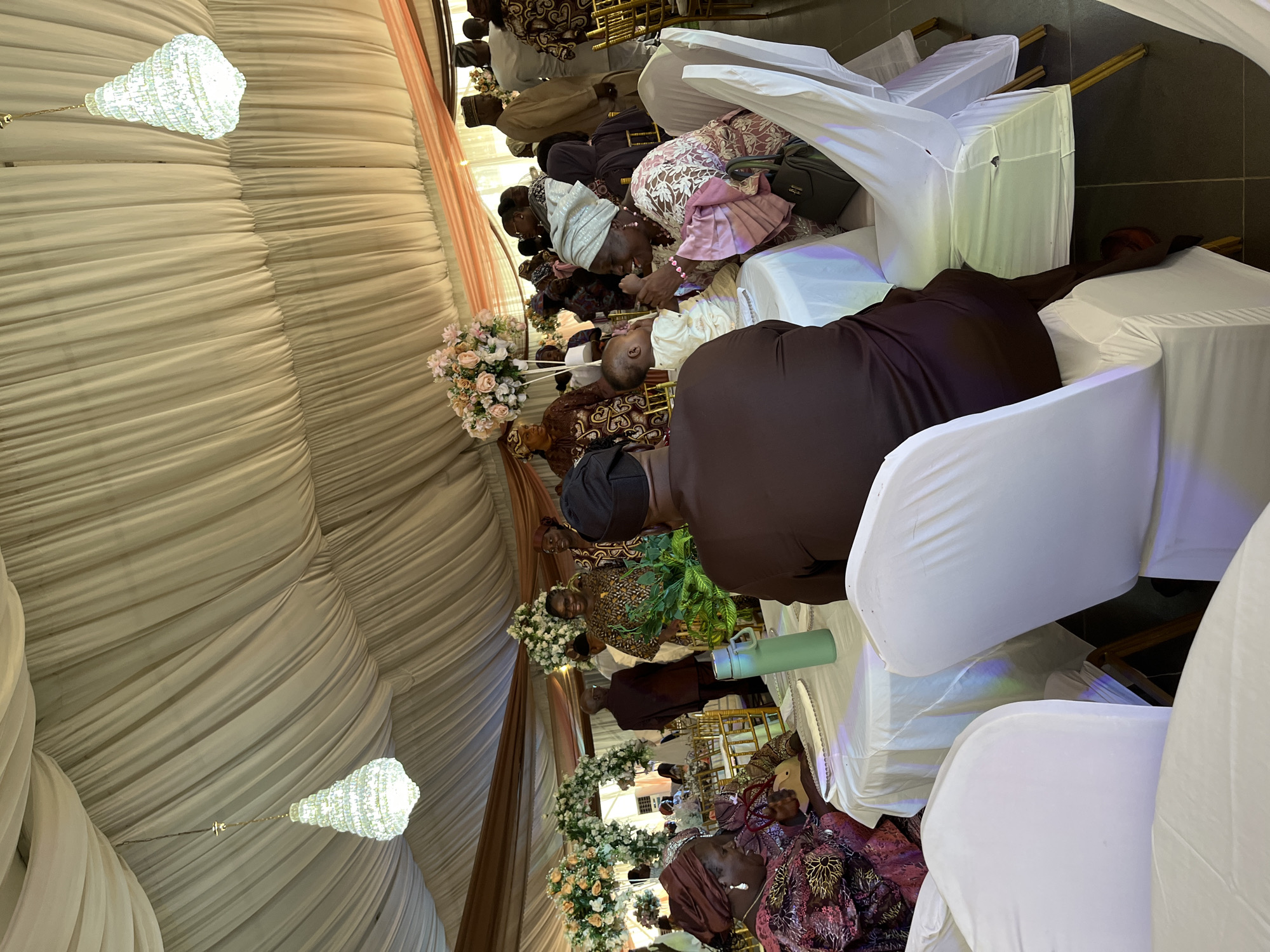 The image shows an event within an interior space likely in Abuja, Nigeria, considering the attire and cultural cues. The setting seems to be a formal gathering, possibly a wedding or celebration, given the tent-like draped fabric covering the ceiling and the chandelier-style lights. Rows of people are seated on white-covered chairs, some interacting with a child. The attire of the attendees is formal and includes traditional Nigerian styles with head coverings and colorful patterns. Flowers are arranged along the walkway, adding to the decorative atmosphere.