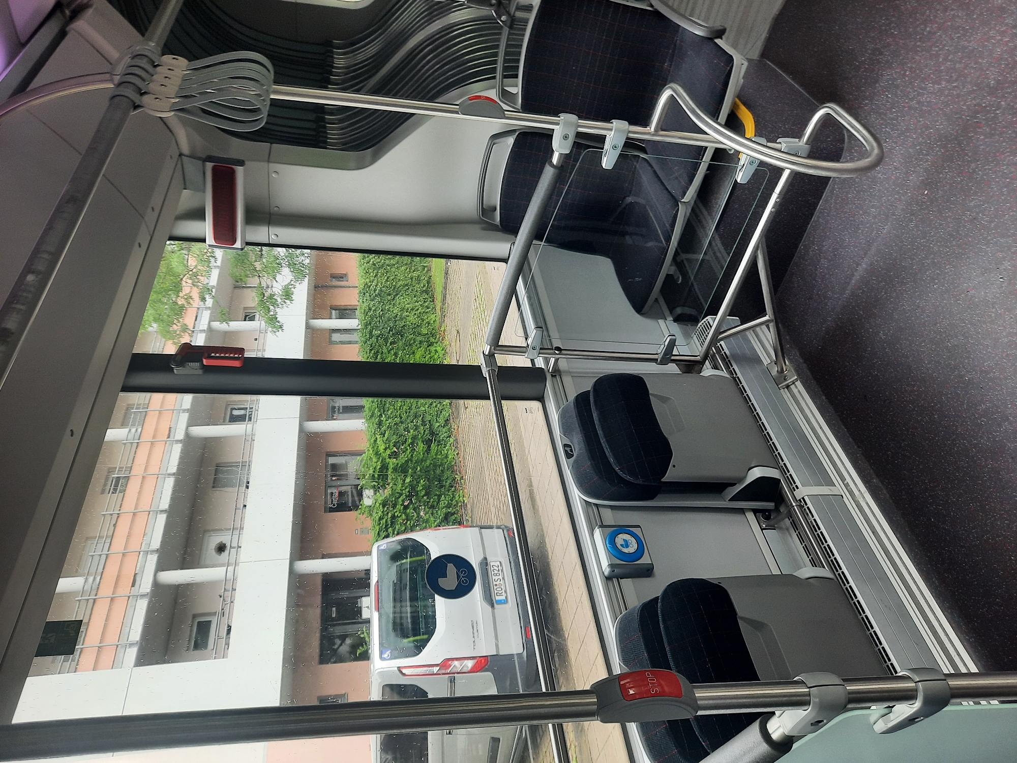 The image shows the interior of a bus in Rosenheim, Germany. Through the windows, a white van with a stroller symbol on the back and a license plate "ROS S 822" is visible, parked outside a building. The bus interior features black and red plaid seats, silver handrails, and a red "STOP" button. The environment outside appears to be an overcast day with rain on the windows, and a green hedge borders the parking area.