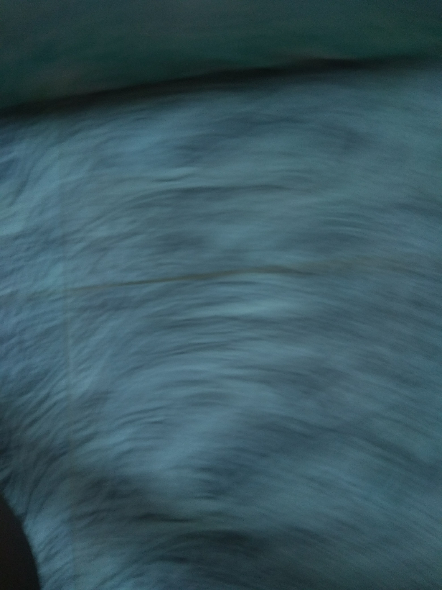 The image is an abstract, low-resolution photograph with a shallow depth of field. It appears to be a close-up of a textured surface, possibly fabric or fur, with a dark area on the left side. The colors are predominantly dark blues and grays, with some lighter streaks that suggest movement or texture. The overall impression is one of blurriness and lack of clear detail. There is no discernible information present in the image that can be used to determine its location within the city of Jalingo, Nigeria. The image does not contain any identifiable people, subjects, objects, actions, interactions, or environmental features that could provide such context.