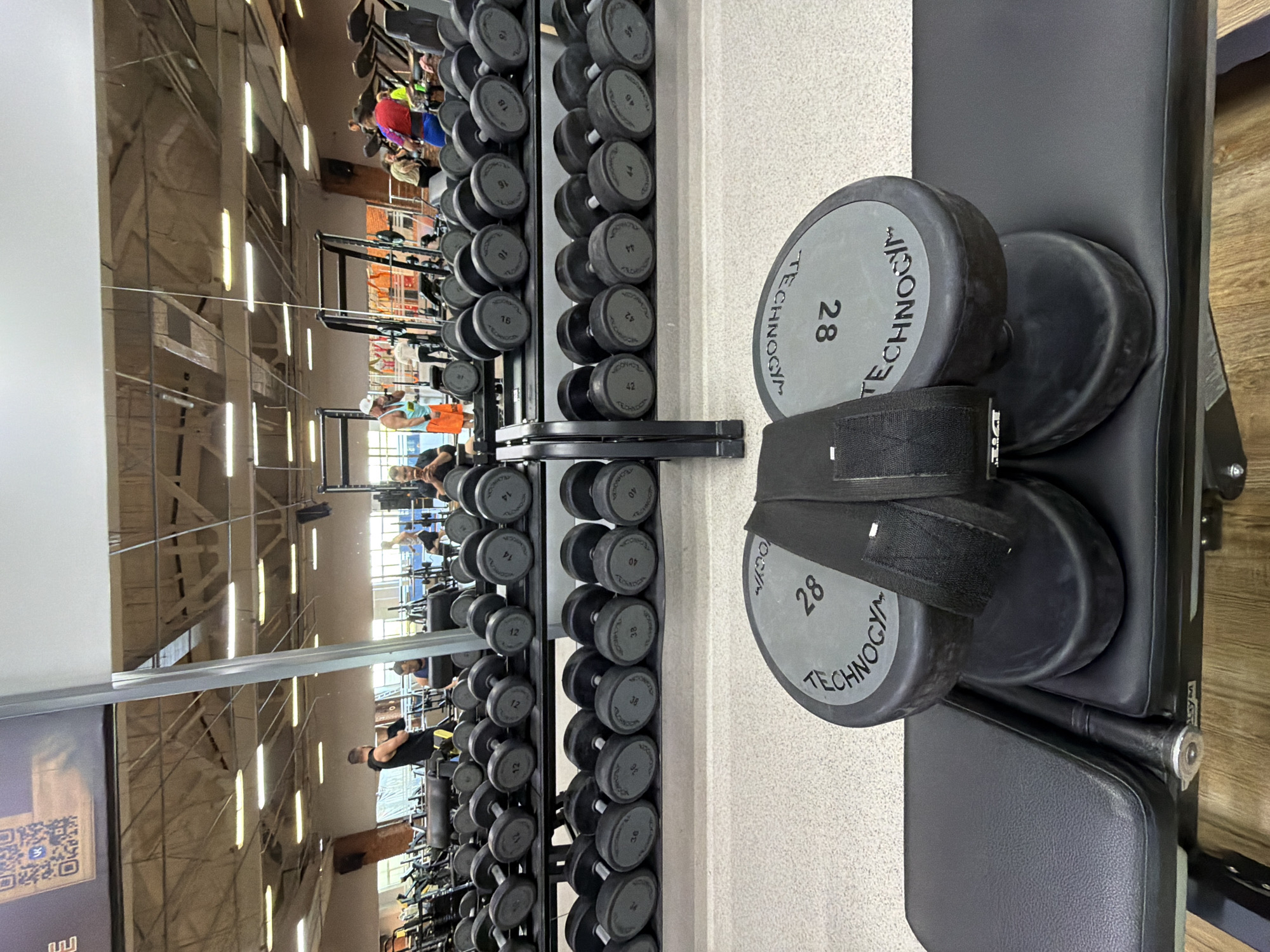 The image depicts the interior of a gym, possibly located in Chaikovsky, Russia, as indicated by the metadata. In the foreground, two large dumbbell weights, each marked with "TECHNOGYM" and "28", are resting on a black weight bench. A black lifting strap is visible around one of the dumbbells. Behind the bench, rows of dumbbells are neatly arranged on black metal racks. The racks extend into the background, with multiple shelves filled with dumbbells of various weights. In the mid-ground and background, several individuals are visible engaged in various activities within the gym. Reflections in a large mirror on the back wall show more of the gym's equipment and people. The environment appears to be a well-lit fitness facility with exposed ceiling structures and fluorescent lighting.