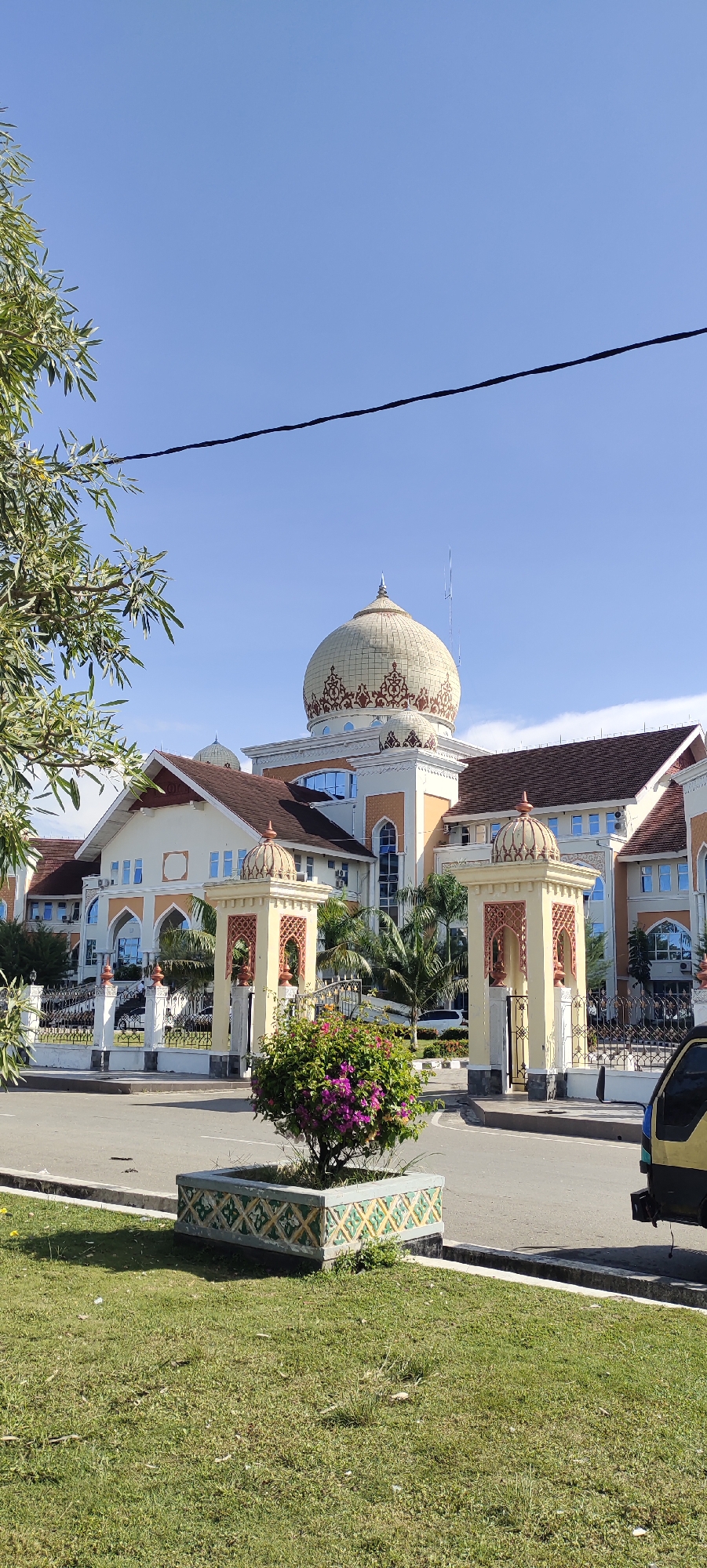 The image captures the exterior of a building with distinct Islamic architectural elements, including a prominent dome and decorative archways, located in Meunasahaluedrienlhoksukon Barat, Indonesia. The building has a light-colored facade with orange accents, a brown tiled roof, and multiple windows. In front of the building, there are ornate yellow gates with Islamic motifs and small domes. A planter box with a flowering bush stands in the foreground, with a neatly mown lawn extending to the street. The sky is clear and blue. A portion of a yellow vehicle is visible on the right side of the image. The scene appears calm and well-maintained, suggesting it might be a public building or a place of worship.