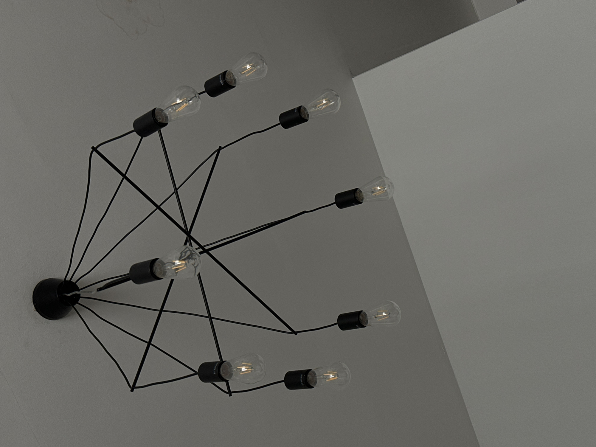 A low-angle, dimly lit shot features a modern, industrial-style light fixture mounted on a white wall. The fixture has a central black hub from which black wires radiate outwards, each terminating in a bulb. The bulbs are clear and display visible filaments. The background consists of a plain, white wall and a portion of a white ceiling at the upper right corner. The lighting is subdued, giving the scene a grayscale appearance. The photograph does not reveal anything indicating the location of Tbilisi, Georgia.