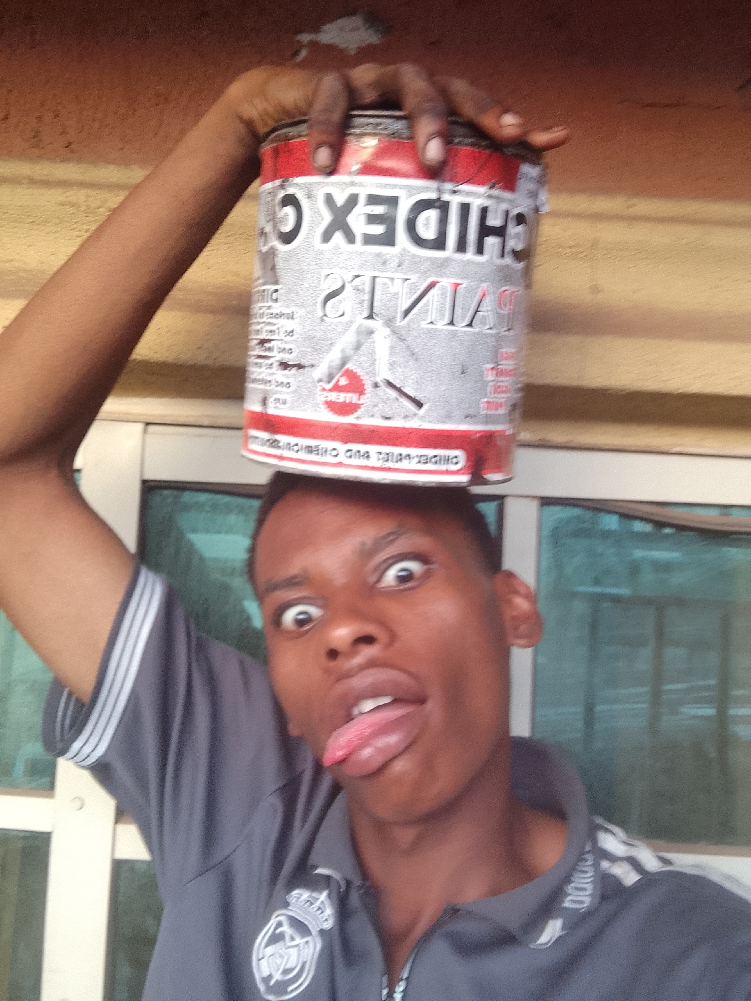 A young Black man is holding a paint can on his head. He is wearing a grey collared shirt and sticking his tongue out with a silly expression. The paint can is red and silver, with the brand name "CHIDEX" visible. The background includes a window with translucent glass and a light brown wall. The location is an undetermined area within the city of Unknown, Unknown.