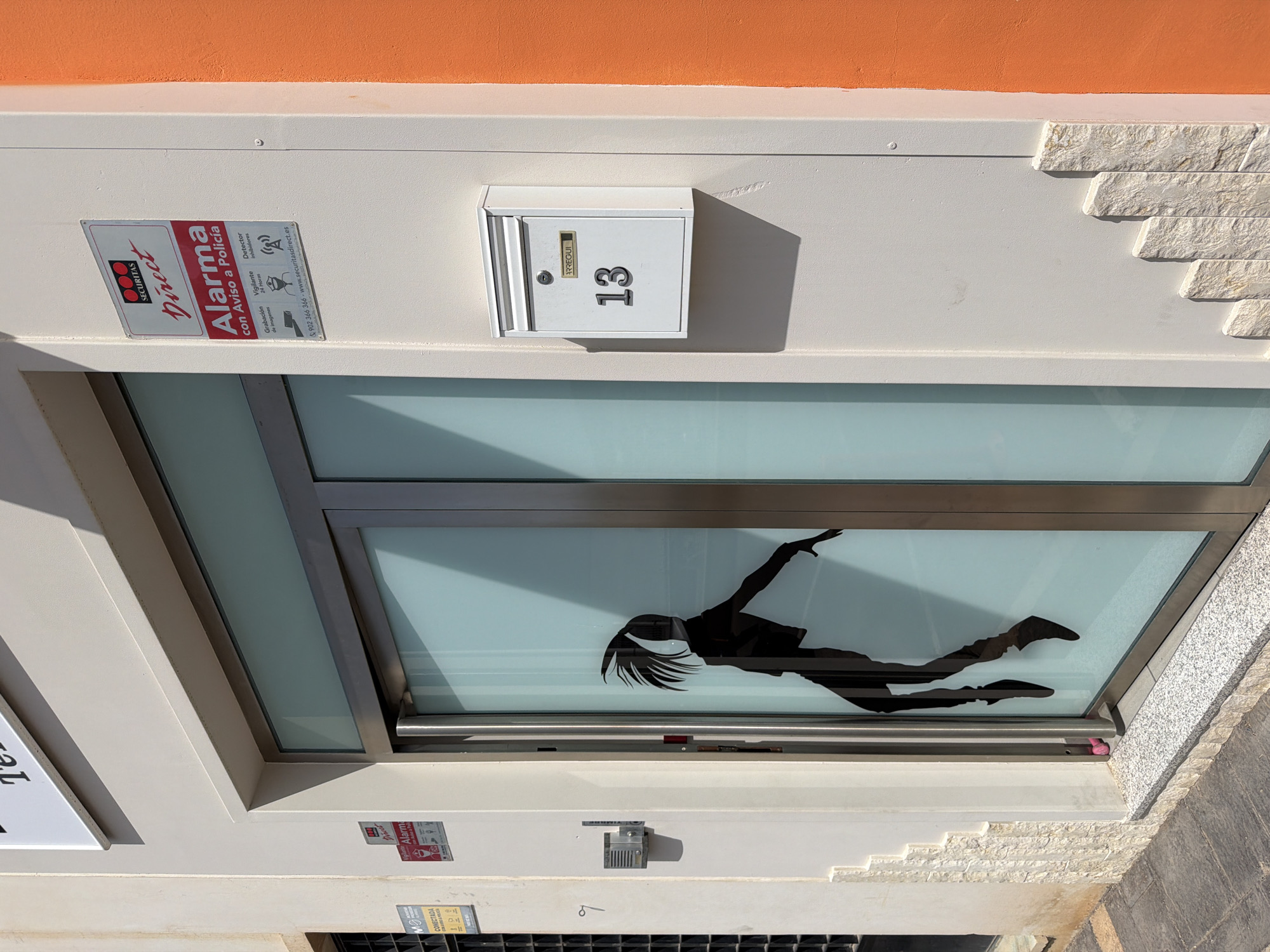 The image shows the facade of a building with an orange accent on the right. A glass door with a silhouette of a person is visible in the center. To the right of the door, there is a white mailbox with the number "13" on it. Above the mailbox, a security sign for "SECURITAS DIRECT" is affixed to the wall. The building appears to be located in a city environment, with cobblestone pavement in the foreground.