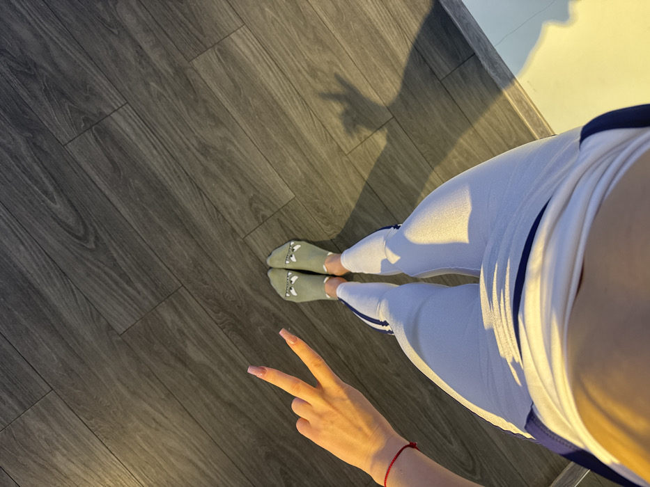 The scene depicts an interior room with dark, diagonally laid wooden floorboards, located in the city of Unknown, Unknown. An individual is observed from an overhead perspective, showing their lower torso, legs, and left arm. They are dressed in white athletic leggings with dark blue vertical stripes along the sides and light green ankle socks adorned with cartoon facial designs. The left hand, displaying pink manicured nails and a thin red string bracelet on the wrist, is raised to form a "peace sign" gesture. Strong, warm directional light from the left illuminates the subject and casts a distinct shadow of the hand gesture onto the floor to the right.
