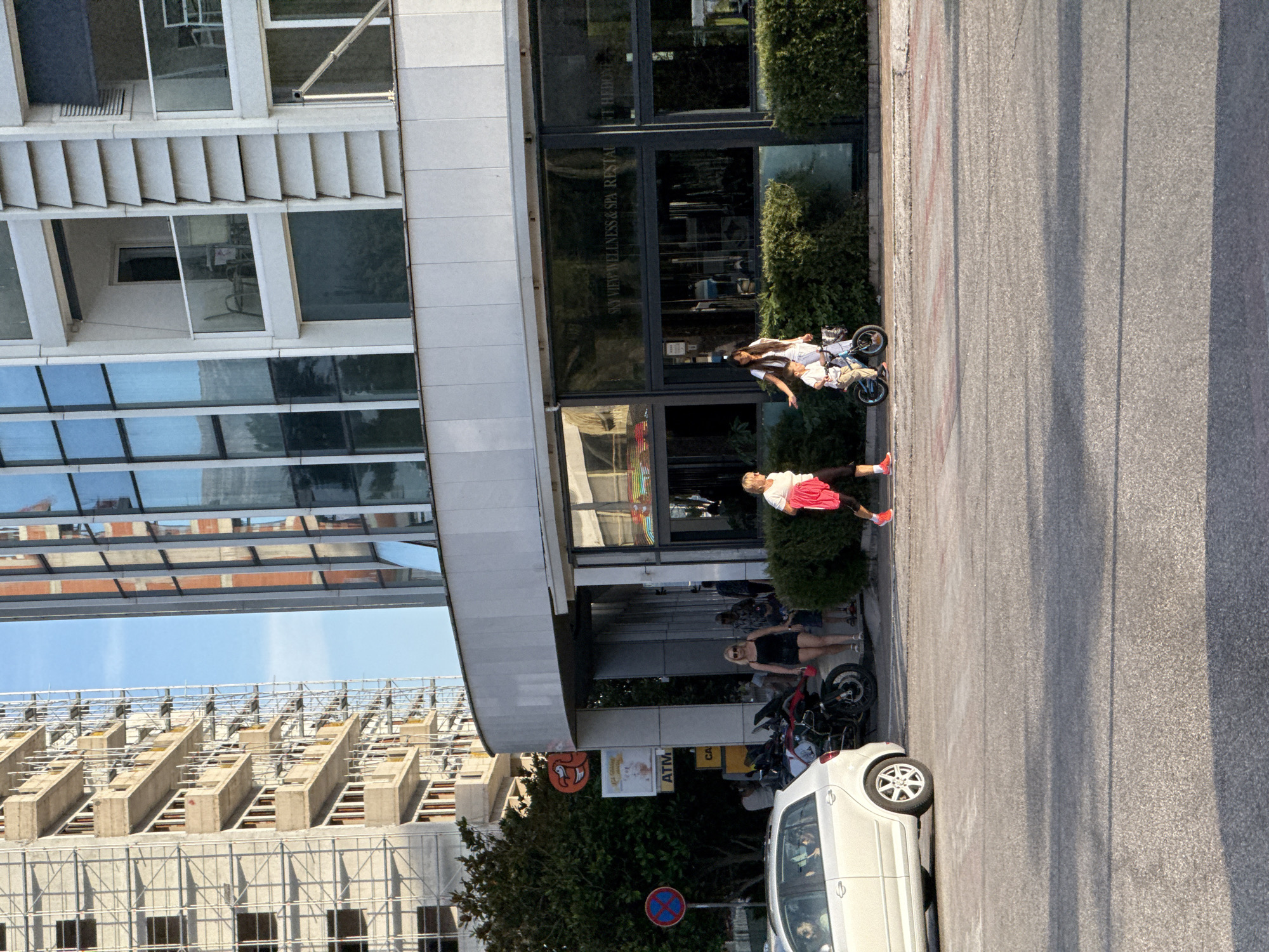 This image depicts a sunny street scene in Budva, Montenegro. The primary subject is a modern, multi-story building on the left, characterized by extensive glass windows and distinctive horizontal architectural elements. The ground floor of this building features a commercial establishment, identifiable by text on its glass facade which reads "SKY VIEW WELLNESS & SPA RESTAURANT".
On the sidewalk in front of this building, several people are visible. In the mid-ground, a woman with dark hair, dressed in a white top, stands beside a young child on a small blue bicycle. The child wears a white shirt and light-colored shorts. Further into the foreground, another woman in a white top and a red skirt or shorts stands near some green bushes. Closer to the camera, a woman with blonde hair, wearing a black top and shorts, is seen near two parked motorcycles.
To the right of the sidewalk, an asphalt road stretches horizontally, exhibiting shadows that suggest either early morning or late afternoon sunlight. A white car is parked on the far right side of the road. Adjacent to the parked motorcycles, an ATM machine is visible with the text "ATM" and partially "CASH". A unique red-orange sign with a stylized face or logo is also present near the ATM. A circular blue traffic sign with a red diagonal line, indicating "no stopping" or "no waiting," is positioned on the sidewalk.
In the background, beyond the main building, a large structure under construction with visible concrete framework and scaffolding is present. The sky above is clear and blue, indicating fair weather. The scene conveys a casual and relaxed ambiance, typical of an urban or resort setting.