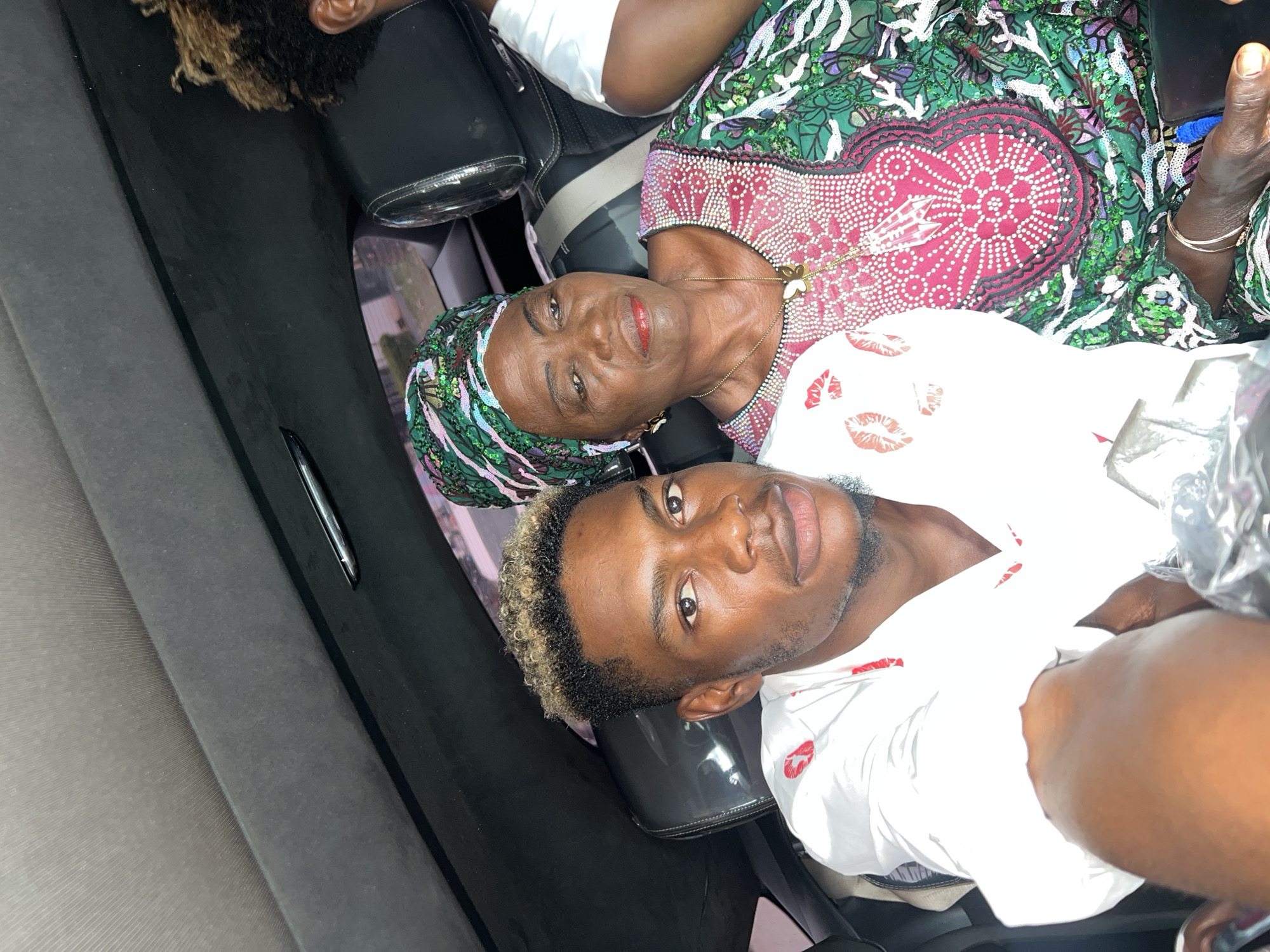 The photograph appears to be taken inside a car, possibly in Igboefon, Nigeria. Two individuals are visible: a younger man with light-tipped hair and a white shirt patterned with red lipstick marks, and an older woman with a green and pink sequined headwrap and matching dress. The woman wears a gold necklace with a flower pendant. The car interior is dark grey. A pen is visible on the car's interior panel. The scene suggests a personal or familial connection between the two subjects.