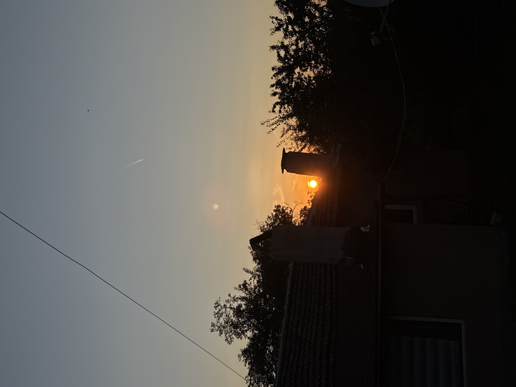 The image depicts a silhouette of a residential house with a tiled roof and a chimney against a dusky sky at sunset. Trees are silhouetted behind the house, and the sun is visible near the horizon, casting a warm orange glow. A thin, dark wire stretches diagonally across the upper portion of the frame. In the distance, a faint white streak, possibly a contrail from an aircraft, is visible in the sky, along with a small, dark object. The scene appears to be a quiet residential area. The location is identified as the city of Sredets, Bulgaria.
