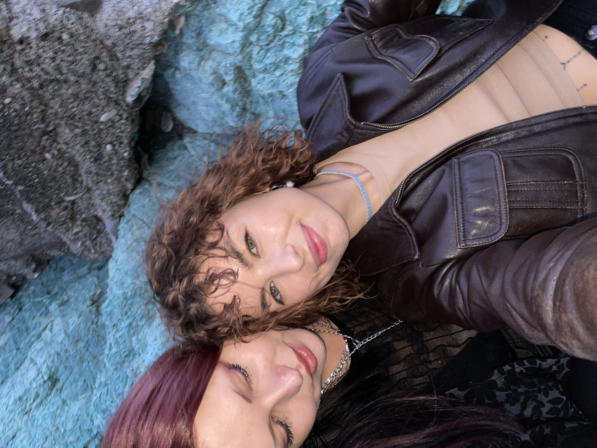 Two women are lying on a bluish-gray rock formation. The woman on top has curly brown hair, green eyes, and is wearing a brown leather jacket, a light beige top, and a blue beaded necklace. The woman below has dark hair with red highlights and is wearing a silver necklace. The location appears to be a rocky area, likely near a beach in Rafailovići, Montenegro.