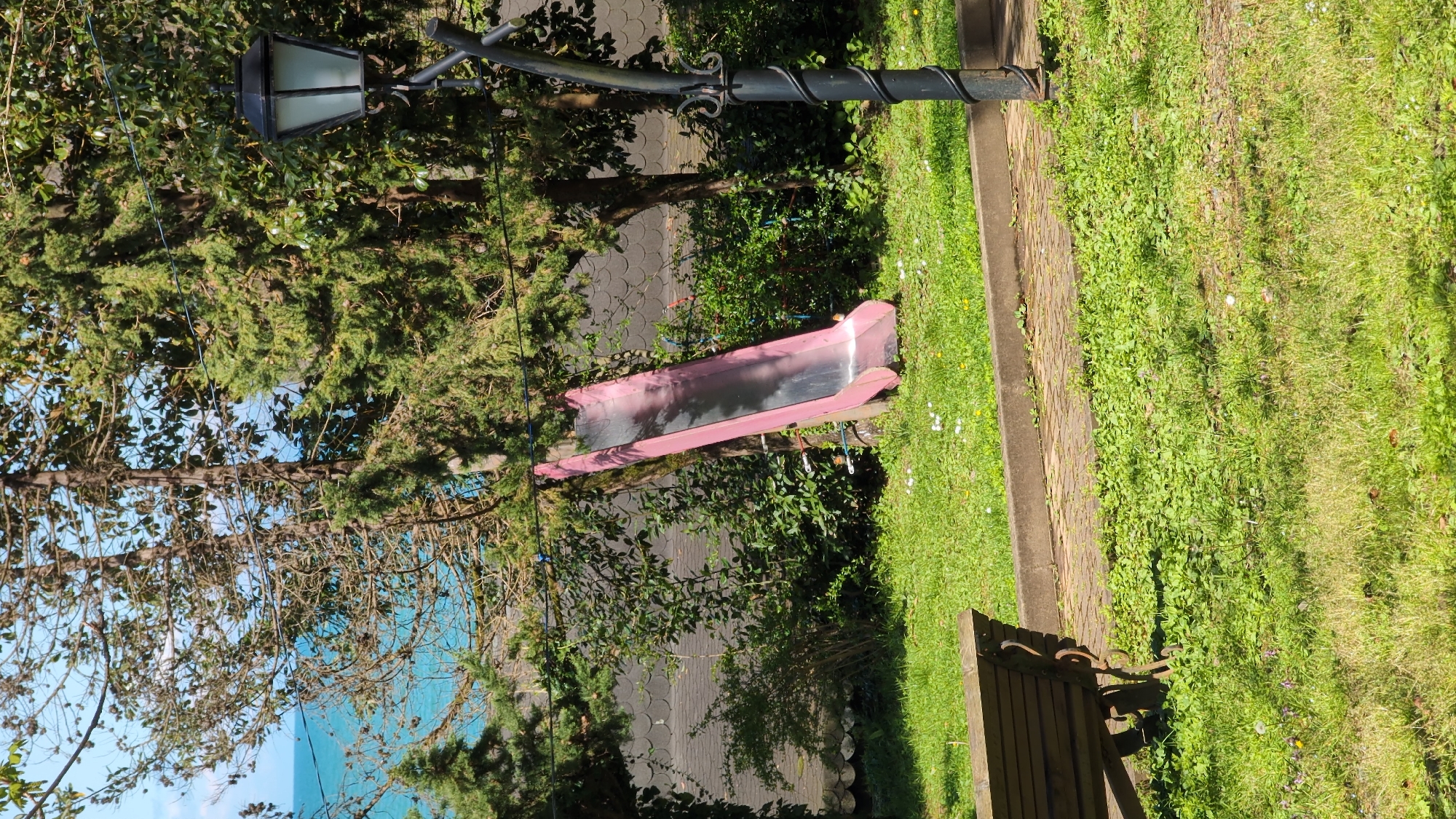 A view of a park in Batumi, Georgia. A pink slide is situated between greenery and trees. A lamppost is nearby, and a bench is in the lower right corner. The ground is covered with grass. A view of the sea is also visible in the distance.