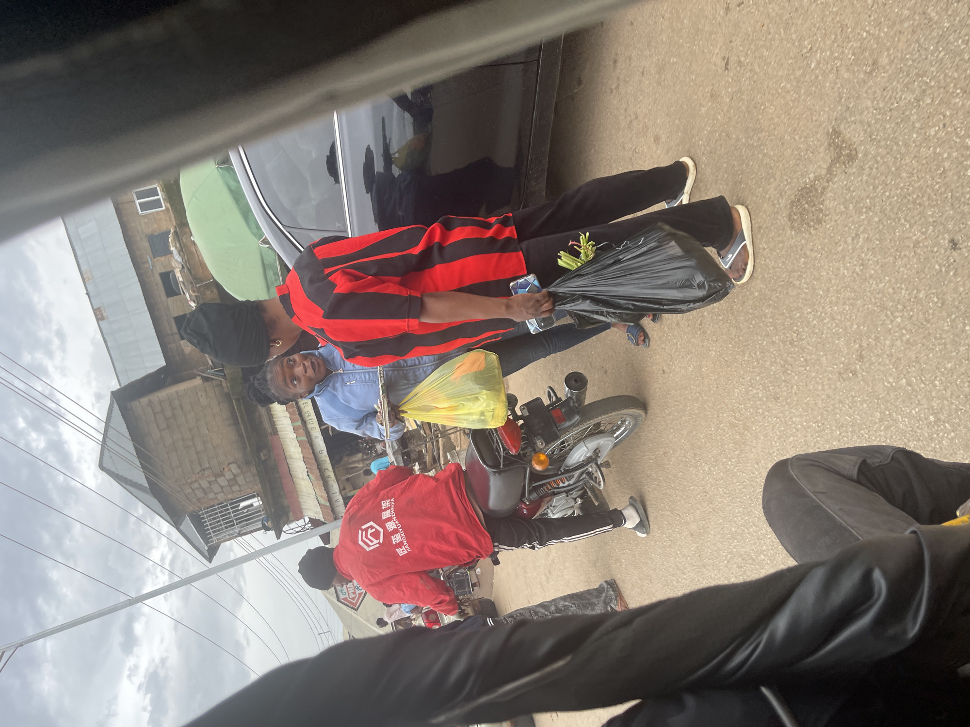 In Gwong, Nigeria, a street scene unfolds under an overcast sky. A motorcyclist, wearing a red jacket with white Chinese characters and black pants, is stopped. Beside him, a woman in a light blue denim jacket and dark pants holds a plate of food and a yellow plastic bag. To her right, another person in a red and black striped shirt and black pants holds a black plastic bag containing green vegetables and a smartphone. A black car is parked behind them. The background shows a brick building with a tin roof, suggesting a commercial or residential area. The ground is paved. The image appears to be taken from a moving vehicle, possibly a trishaw, due to the blurred foreground elements.