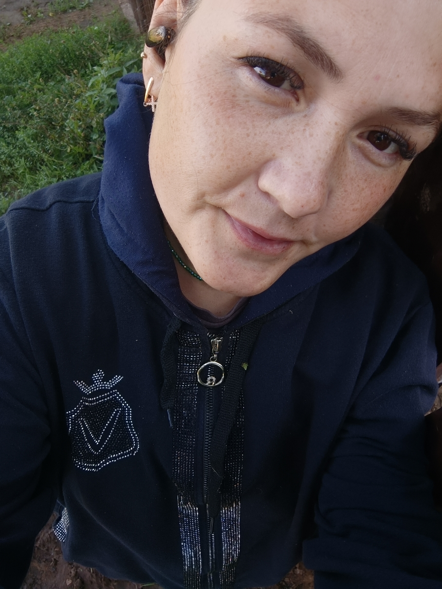 The provided image is a close-up, high-angle, outdoor photograph of a young woman's face and upper body. She is wearing a dark blue hooded jacket with a zipper and decorative embellishments on the left chest, appearing to be a crown above a shield-like shape filled with small reflective elements. A dark green beaded necklace is visible around her neck. She has fair skin with numerous freckles across her nose and cheeks. Her eyes are dark brown, and she has dark eyelashes and eyebrows. She is wearing multiple earrings in her left ear: a small stud, a gold hoop, and a cross-shaped earring. The background consists of green foliage and what appears to be dry earth or mud. There is no visible indication of the location being the city of Karsha, Russia.