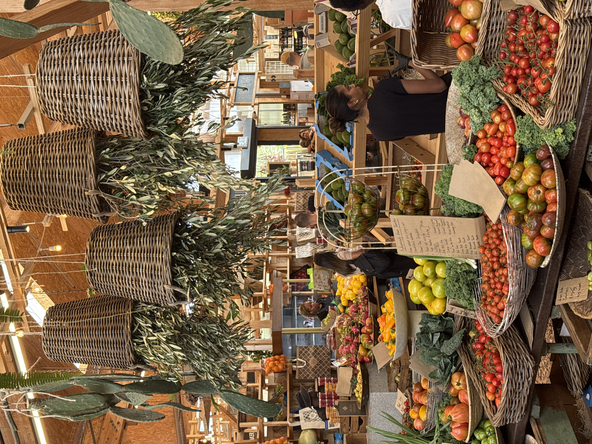 The image shows an indoor marketplace or farm stand, possibly located in Cape Town, South Africa, based on the labels on produce. Hanging overhead are woven baskets containing olive branches and cactus plants. Below, customers browse produce including tomatoes, peppers, and avocados. The structure of the stand is primarily wood.